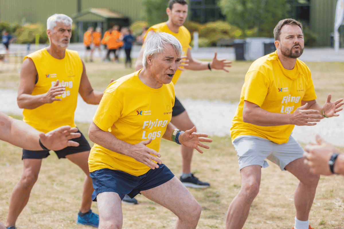 Outdoor team-building event with participants wearing matching blue shirts, taking part in inflatable obstacle courses and games, smiling and cheering, with the words 'ENTER A TEAM' in large bold white and green text across the image.