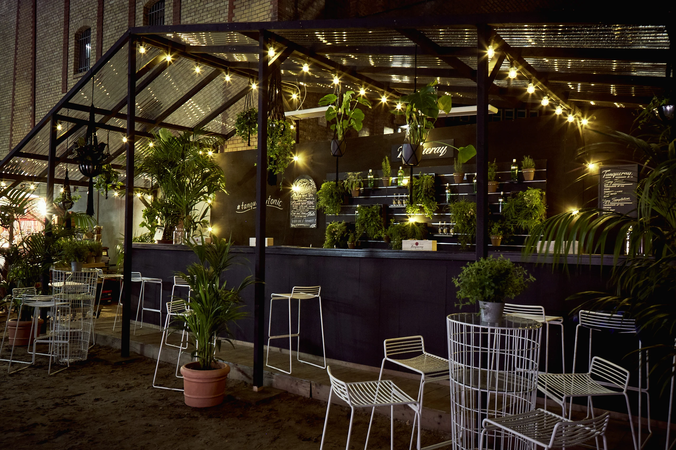 Greenhouse-style Tanqueray bar at DIAGEO BCB17 with string lights, hanging plants, white wire chairs and chalkboard menus at night.