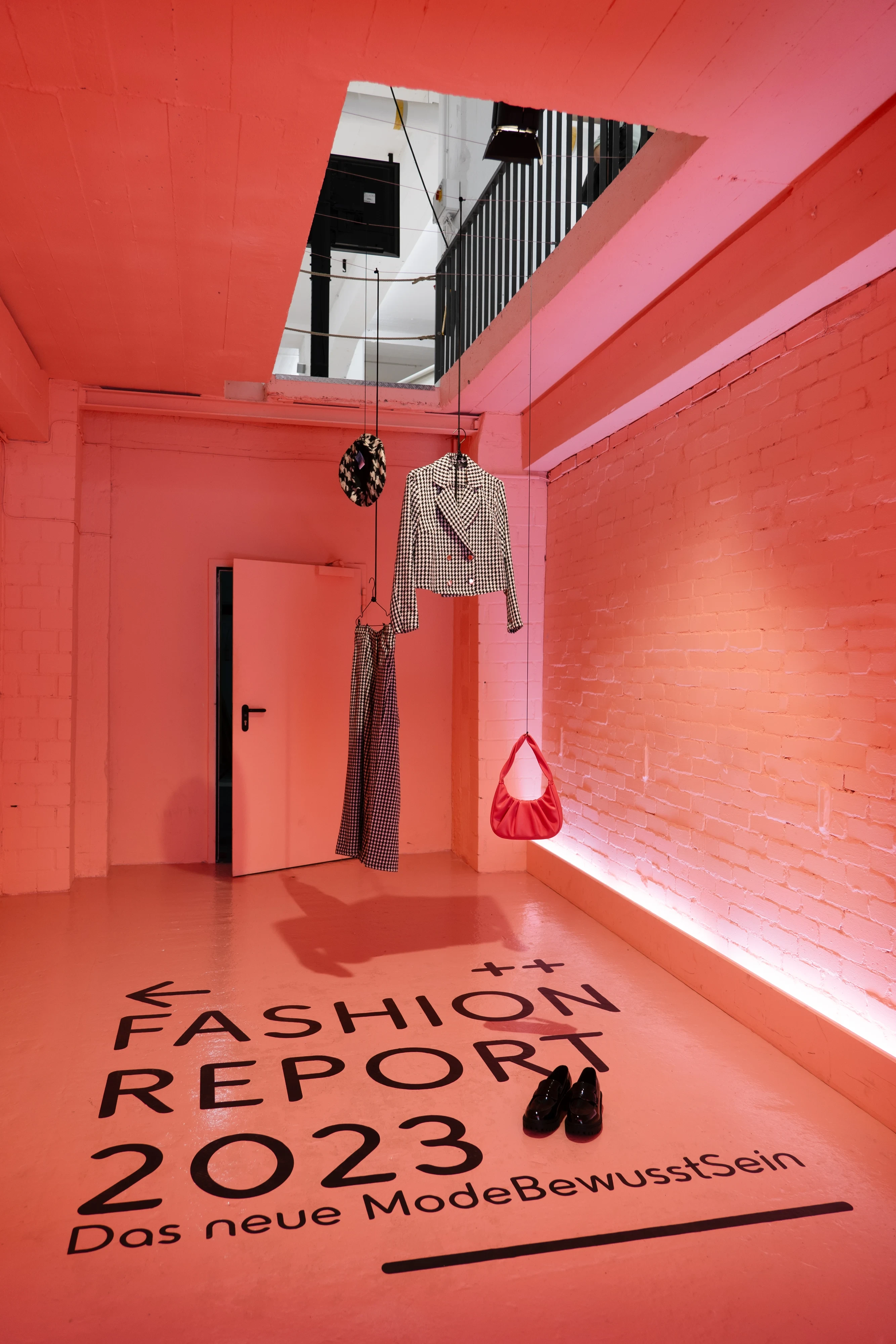 Coral-lit brick room with Fashion Report 2023 floor lettering and hanging outfits.