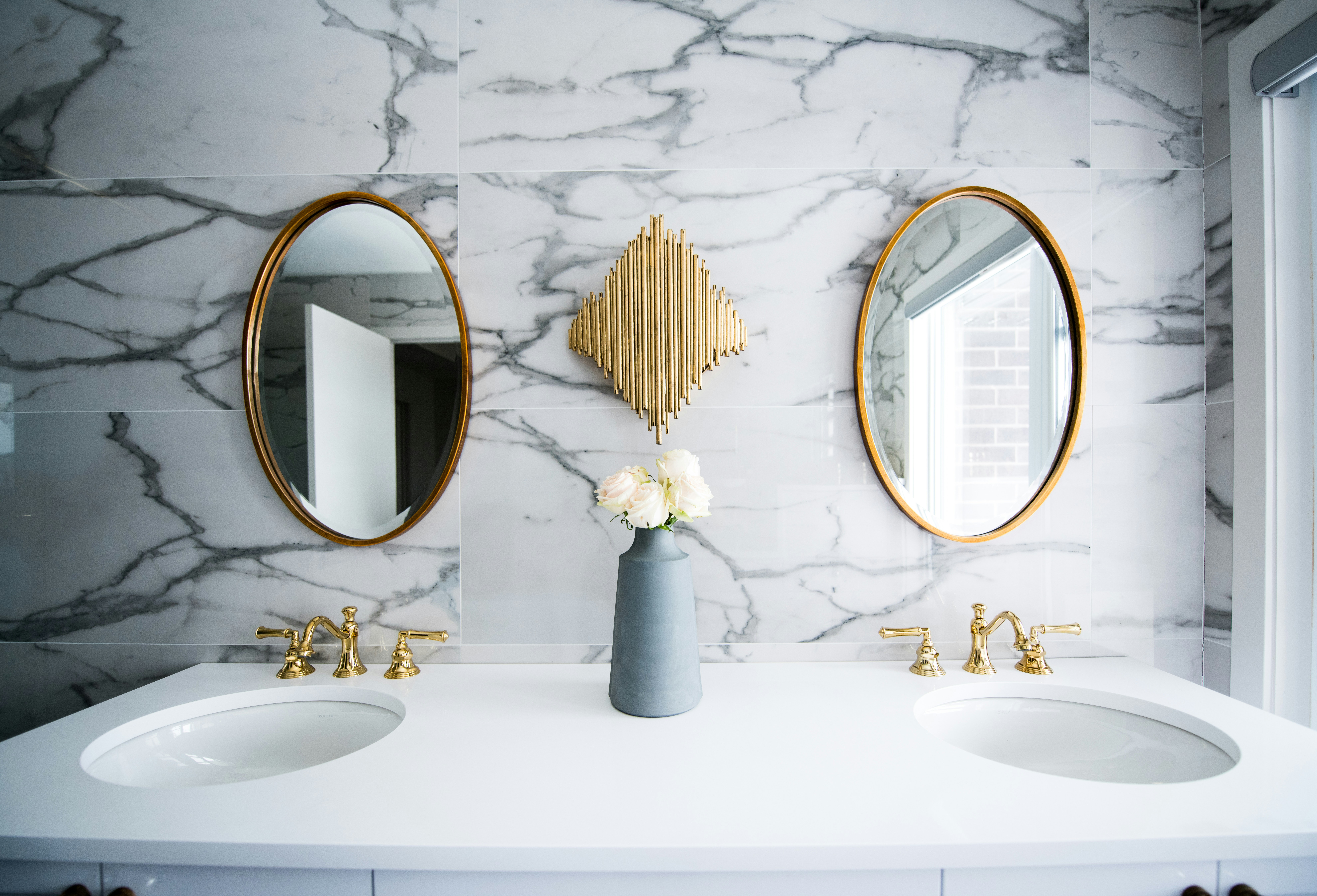 A clean modern bathroom with a sink, marble walls and 2 oval mirros with gold accents