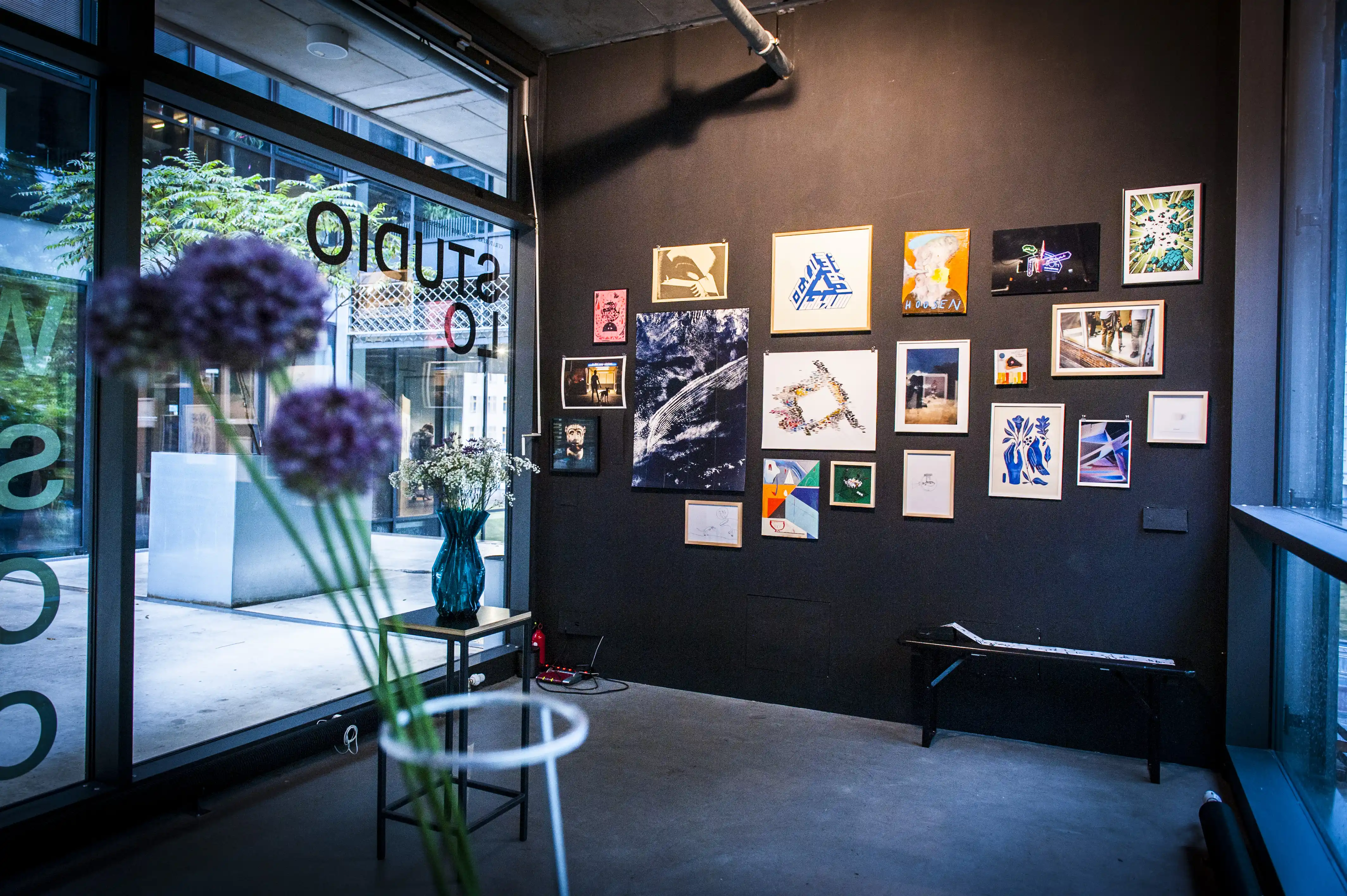 Inside the STUDIOLO gallery with a salon-style wall of framed photographs and artworks on black, a black bench and purple allium flowers in the foreground.