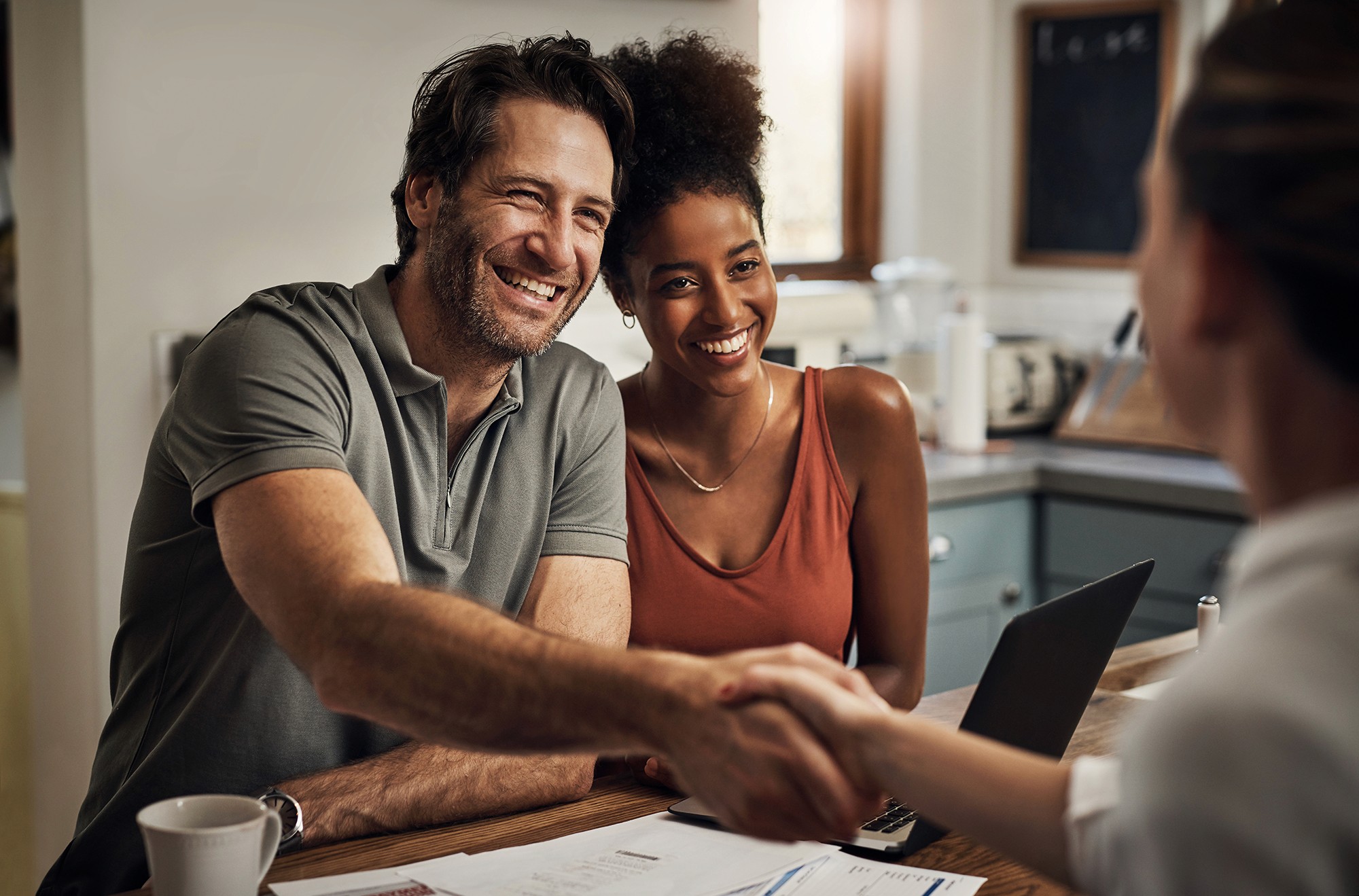 a happy couple shaking hands with another person while sitting at a table in a kitchen. A mug, some papers, and a laptop are on the table. 