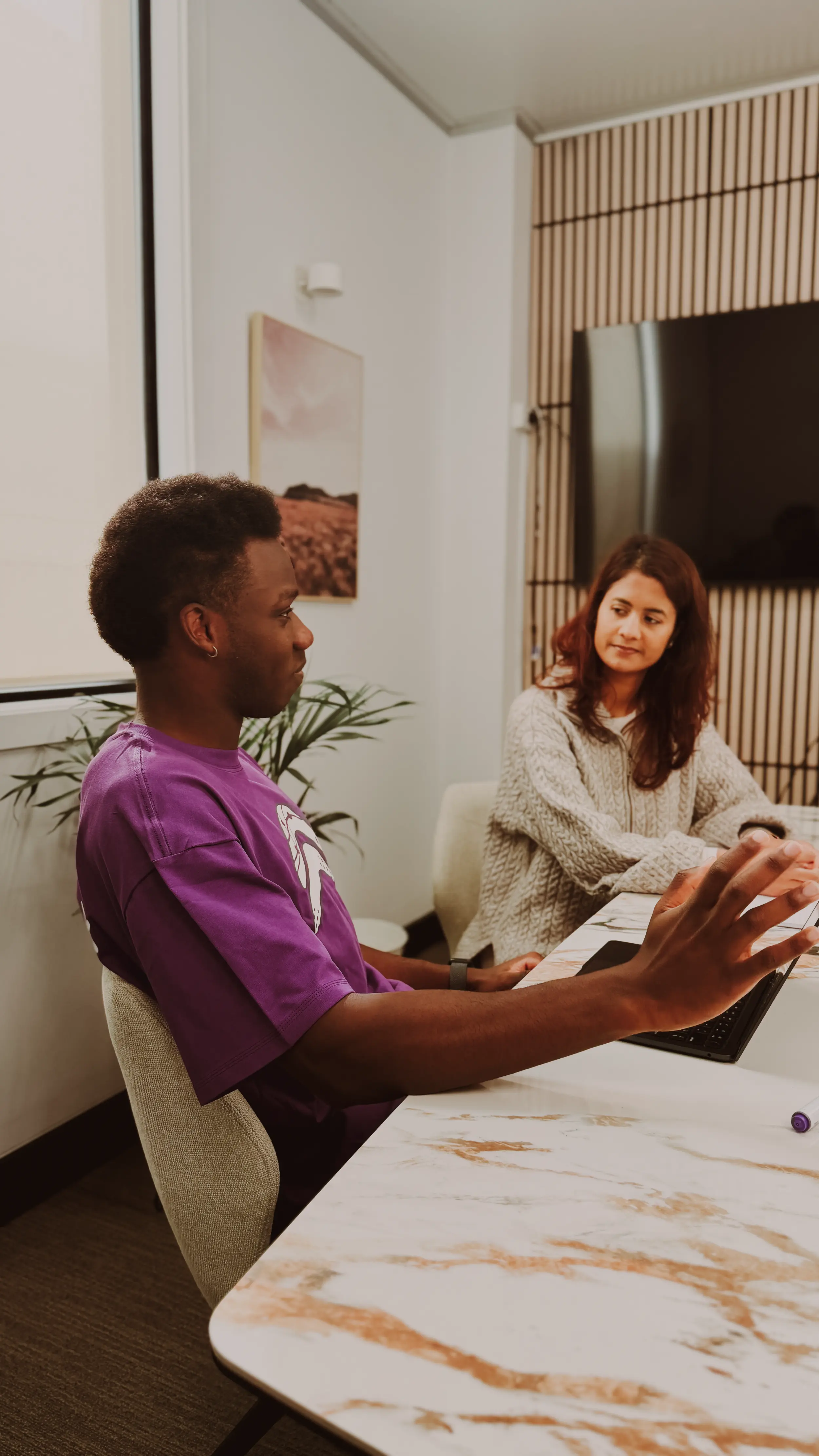 Two people are engaged in a discussion in a modern conference room, featuring a large marble table, a laptop, and a wall-mounted television.