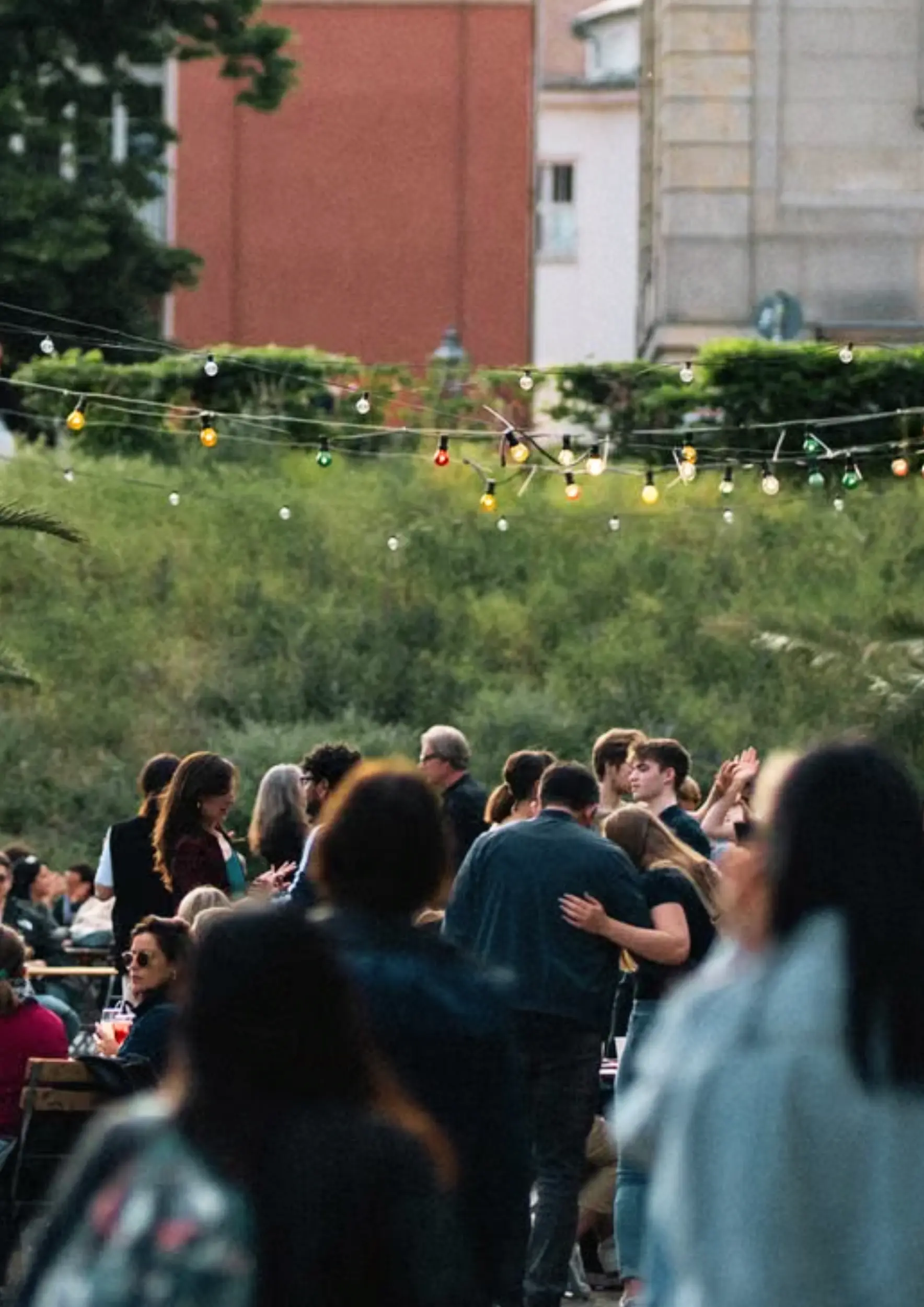 an outdoor party; a group of people are visible through a mellow crowd; two men embrace in conversation under colored string lights