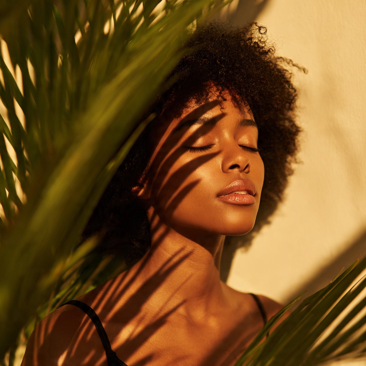 Woman with curly afro hair rests under palm leaf shadows.