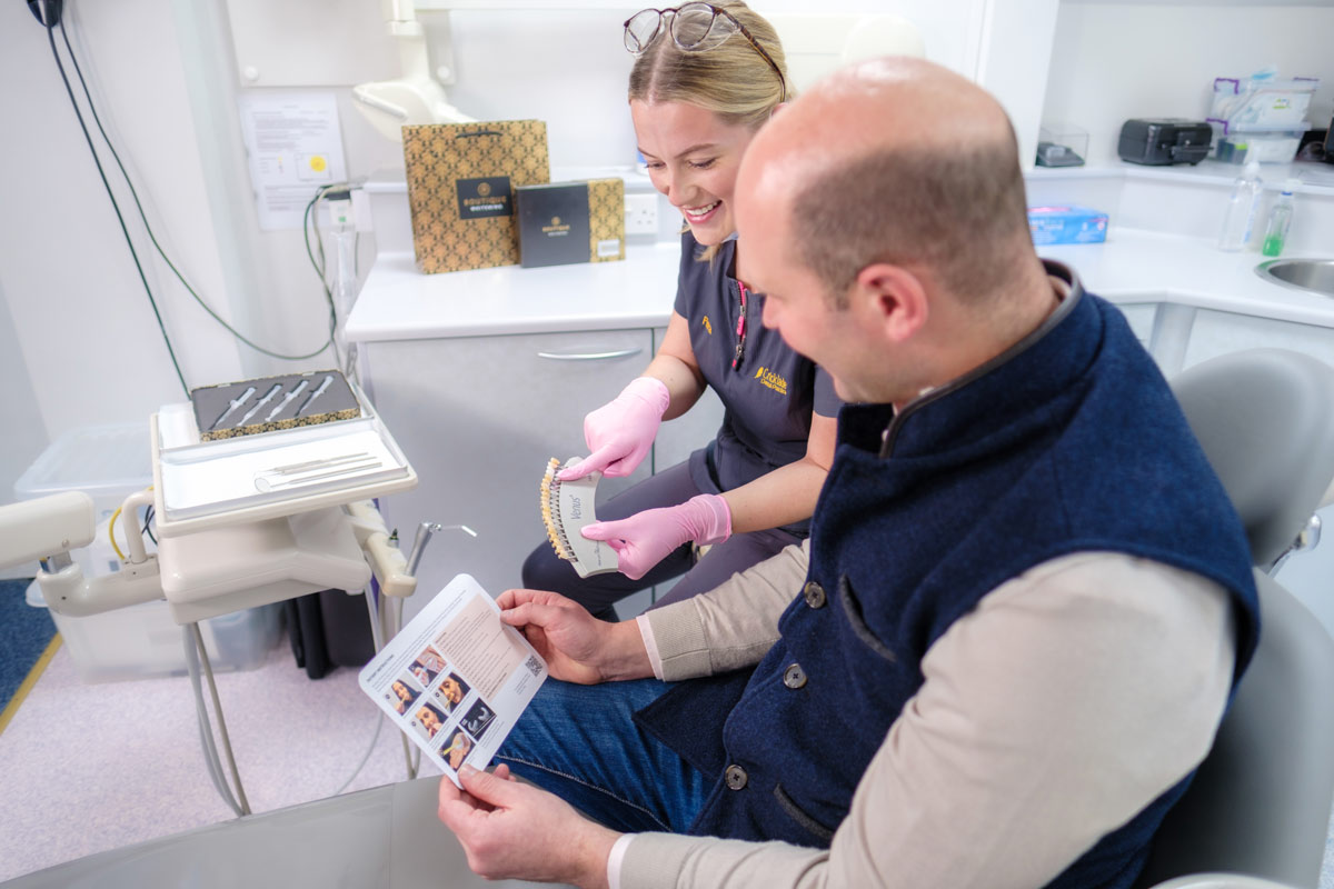 A candid shot of Flora, an Associate Dentist, consulting with a male patient in the dental chair about tooth whitening. She is wearing pink gloves and holding a tooth shade guide for comparison, while the patient holds an instruction leaflet. Gold and black "Boutique Whitening" product kits are displayed on the counter in the background.