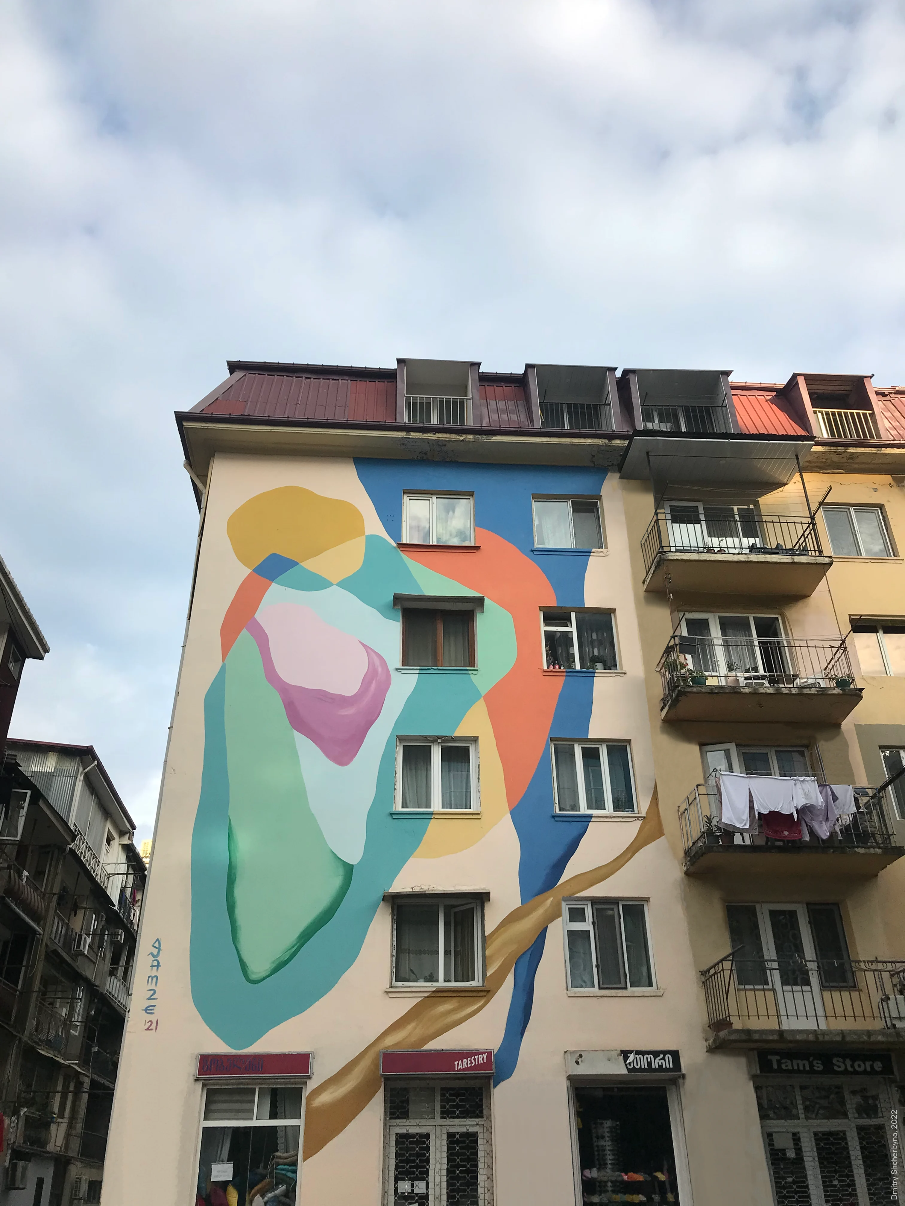A mural on the side of an apartment building depicts two abstract figures in an embrace rendered in soft, overlapping shapes of blue, coral, mint green, yellow and pink.