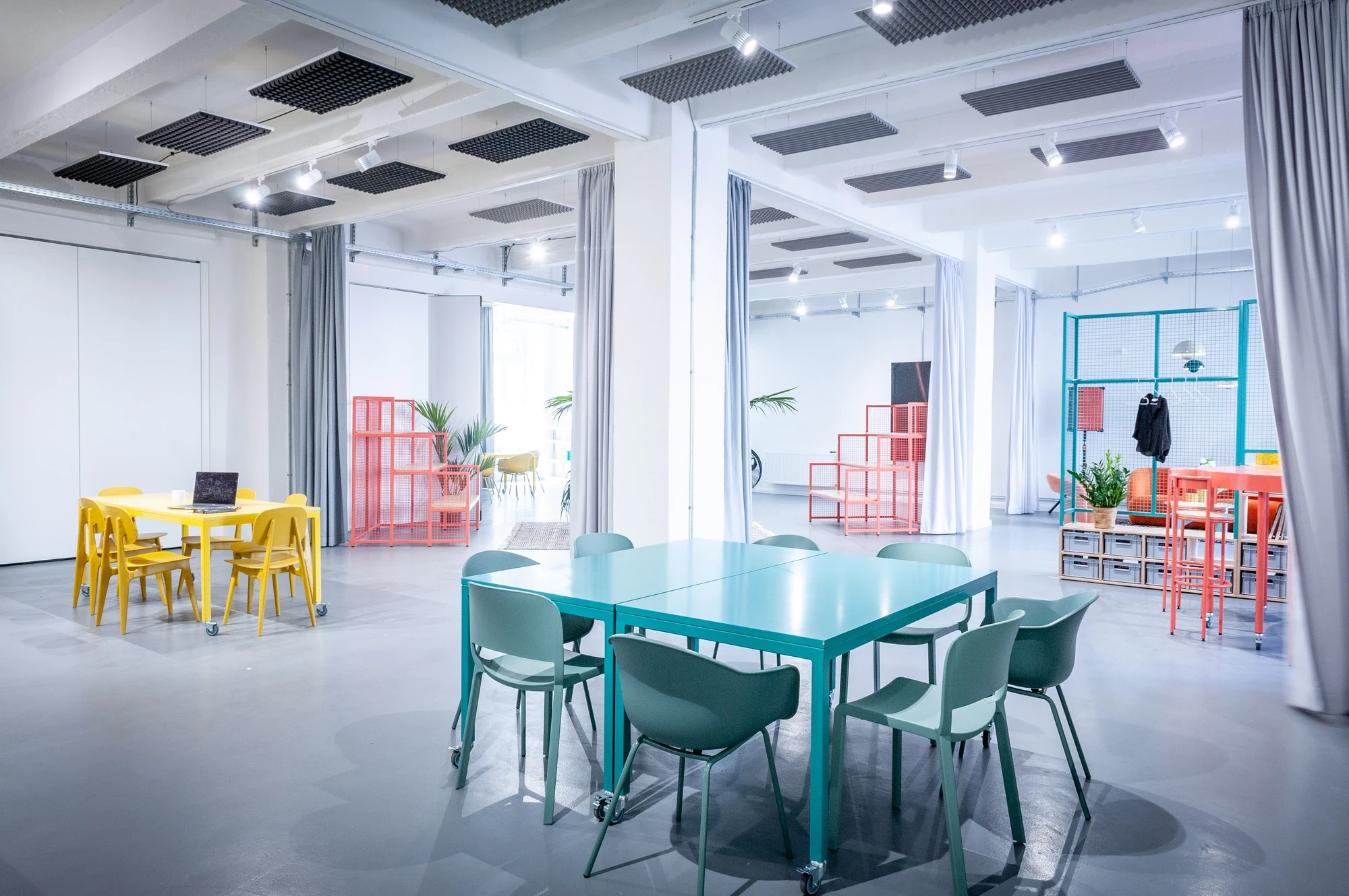 Bright open coworking space with yellow, teal and coral colour-blocked tables, modular metal grid dividers and floor-to-ceiling curtains.