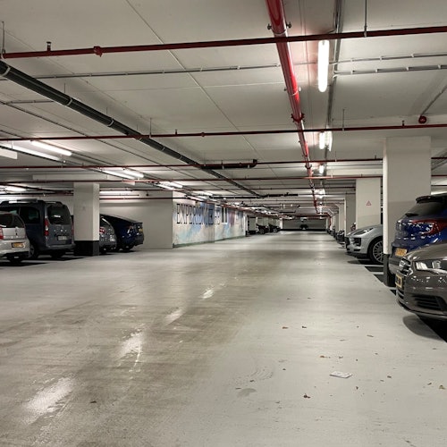 地下停车场内停泊着多辆汽车，白色的墙壁，天花板上有红色的管道和悬挂的荧光灯。