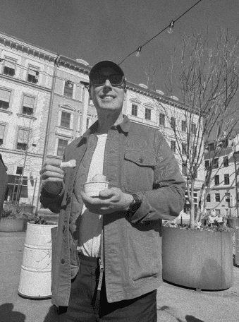 Black-and-white photo of a person standing outdoors in a city square, holding a small cup and spoon.