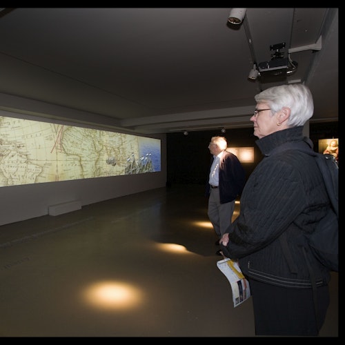Two people in a dimly lit exhibit room looking at a large historical map projected on a wall.
