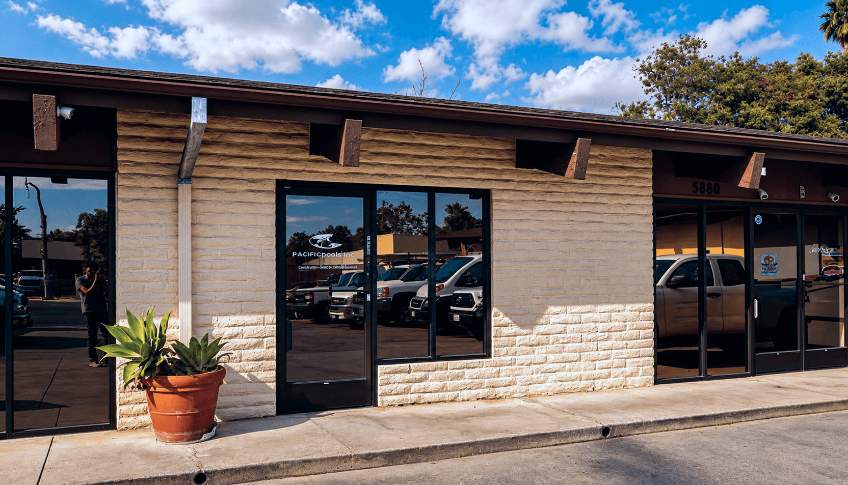 Street-level facade of a single-story commercial retail building with textured tan brick, dark bronze-framed glass storefronts, brown canopy overhangs, and a potted plant at the entrance, with parking visible through the reflective glass.