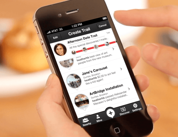 A person's hands hold a black iPhone displaying the AT&T 3G app. The screen shows "Create Trail" at the top, with three profile pictures below: "Afternoon Date Trail," "Jane's Carousel," and "ArtBridge Installation." Each profile includes a circular image and a short description. The time displayed is 1:22 PM. The background is blurred, showing an orange surface.