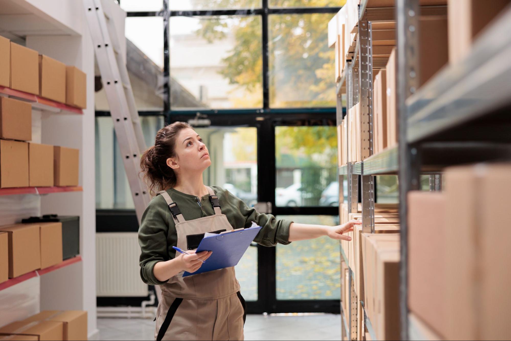 A woman in overalls holds a clipboard, inspecting cardboard boxes  on a heavy duty steel open rack in a warehouse. Large windows in the background let in soft natural light.