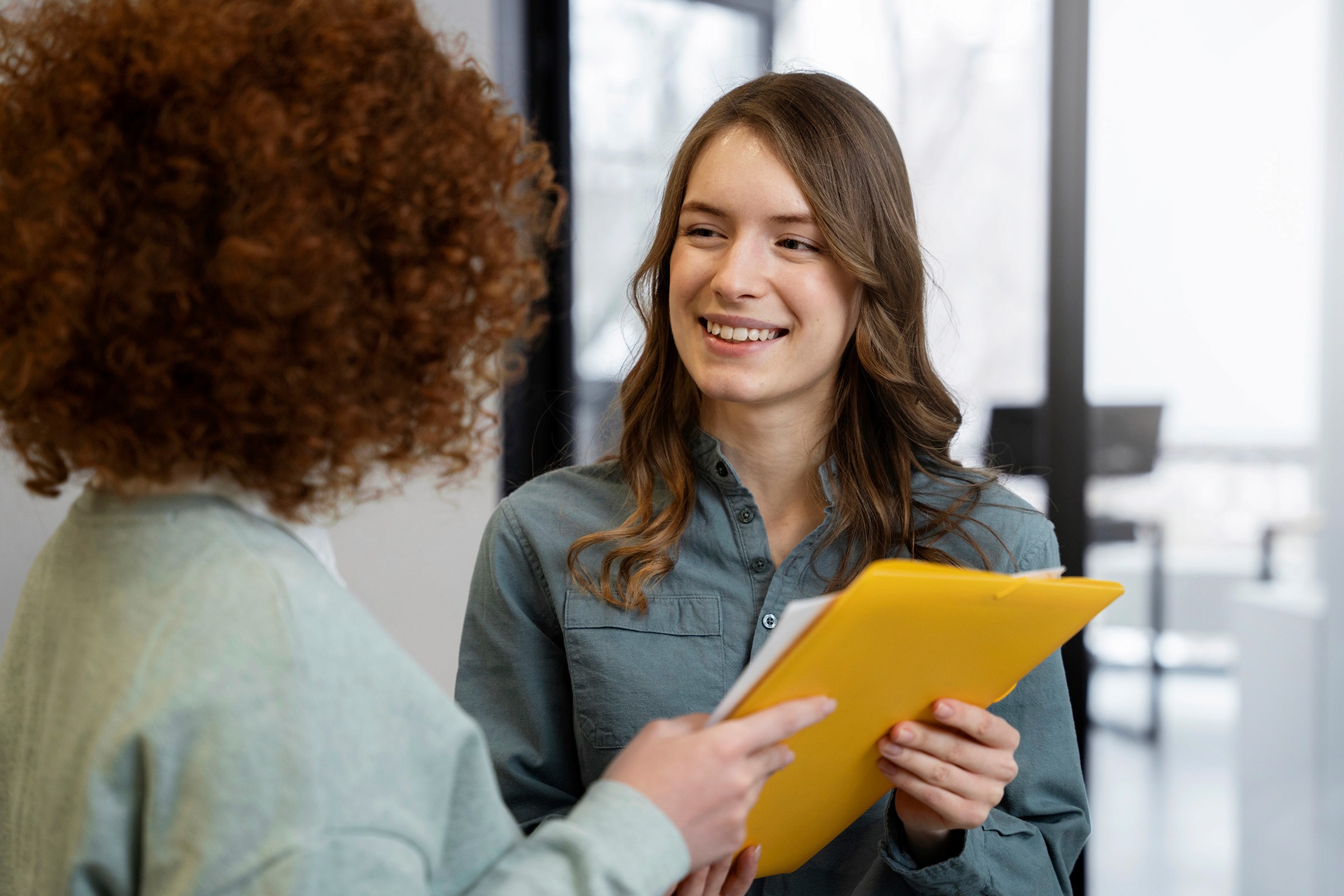 A young woman smiling while holding a yellow folder and engaging in a conversation with a colleague in an office setting