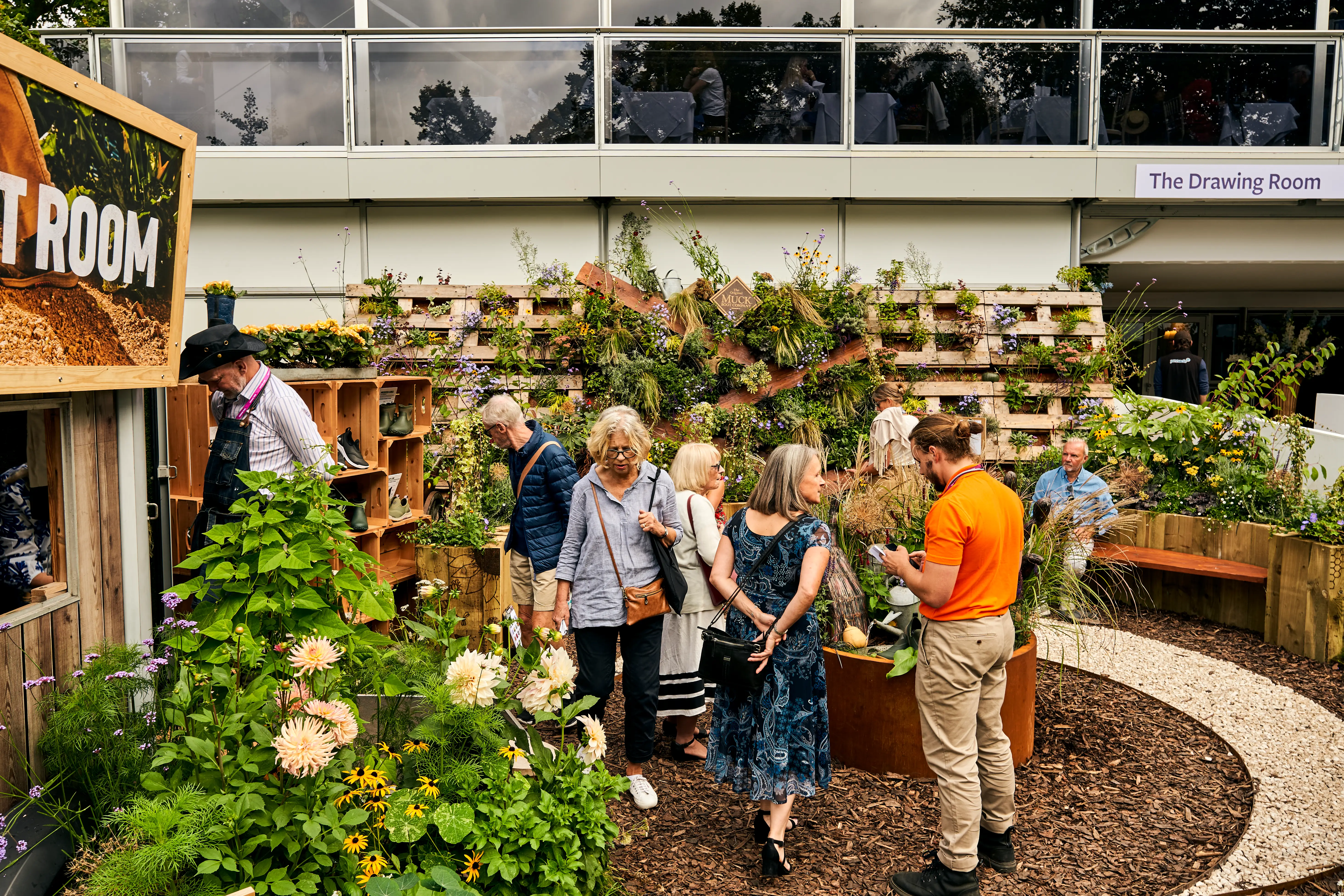 People interacting in a garden area with plants and a building in the background. Bright, sunny atmosphere.