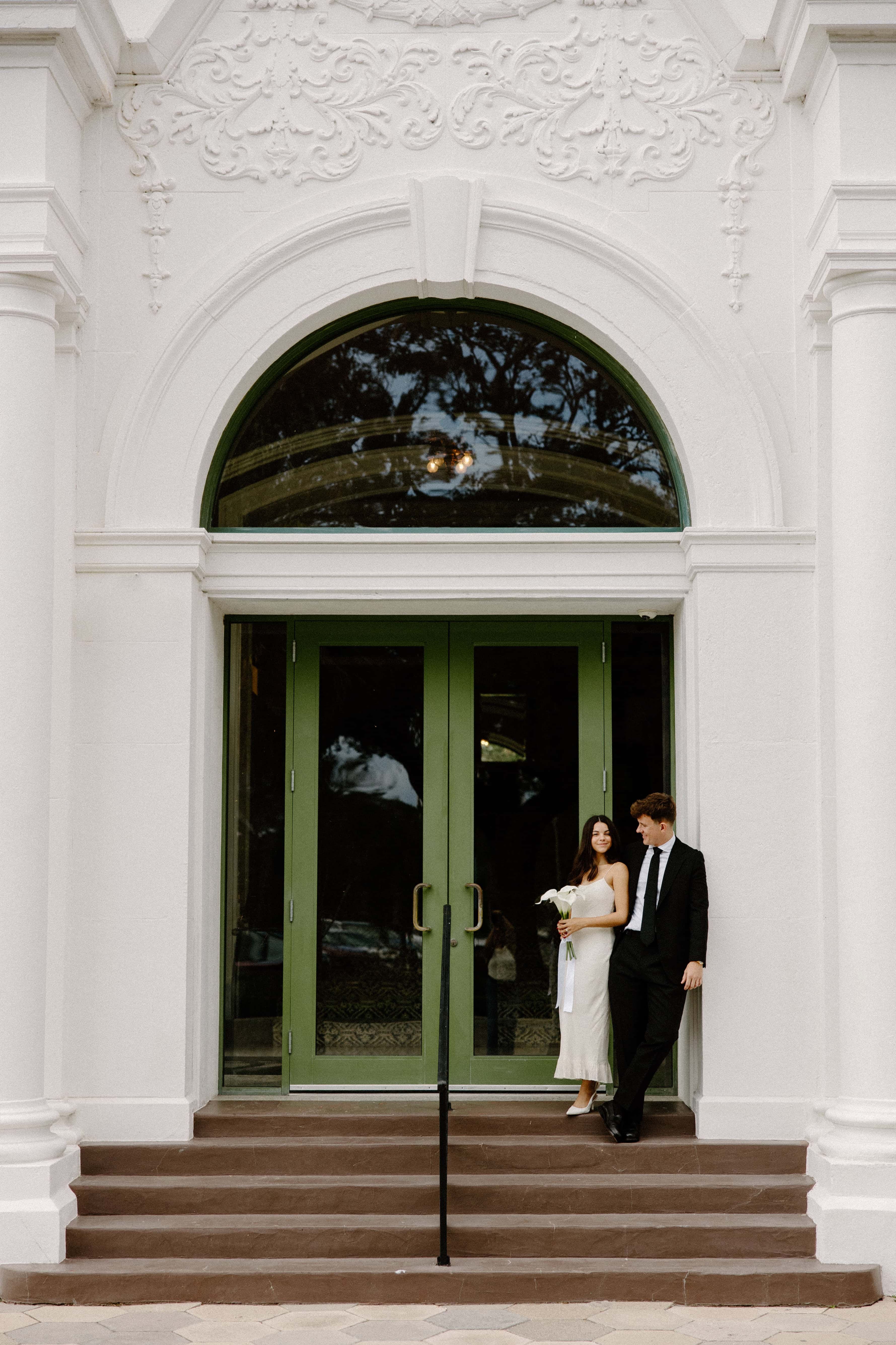A photo of a wedding couple standing on the steps in front of a large, white building with a green double door and an arched window above it.