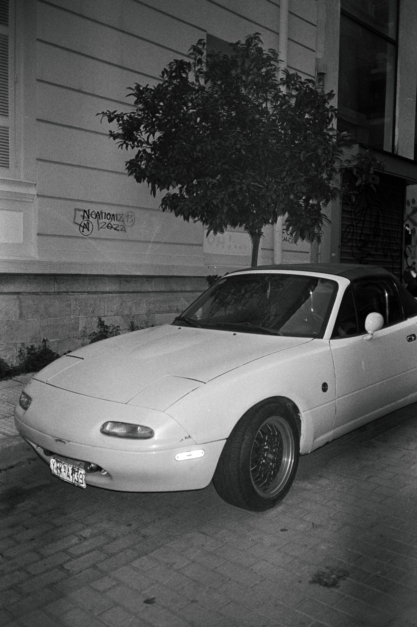 A classic white convertible sports car is parked on a cobblestone street beside a building, with a small tree and graffiti on the wall in the background.