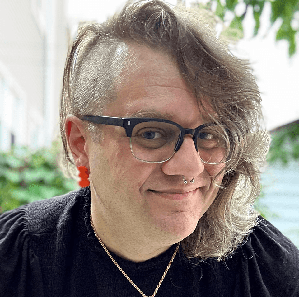 A portrait of Jane wearing a dark shirt and orange earrings in an outdoor setting. Jane has glasses, a nose ring, and long hair with a side shave