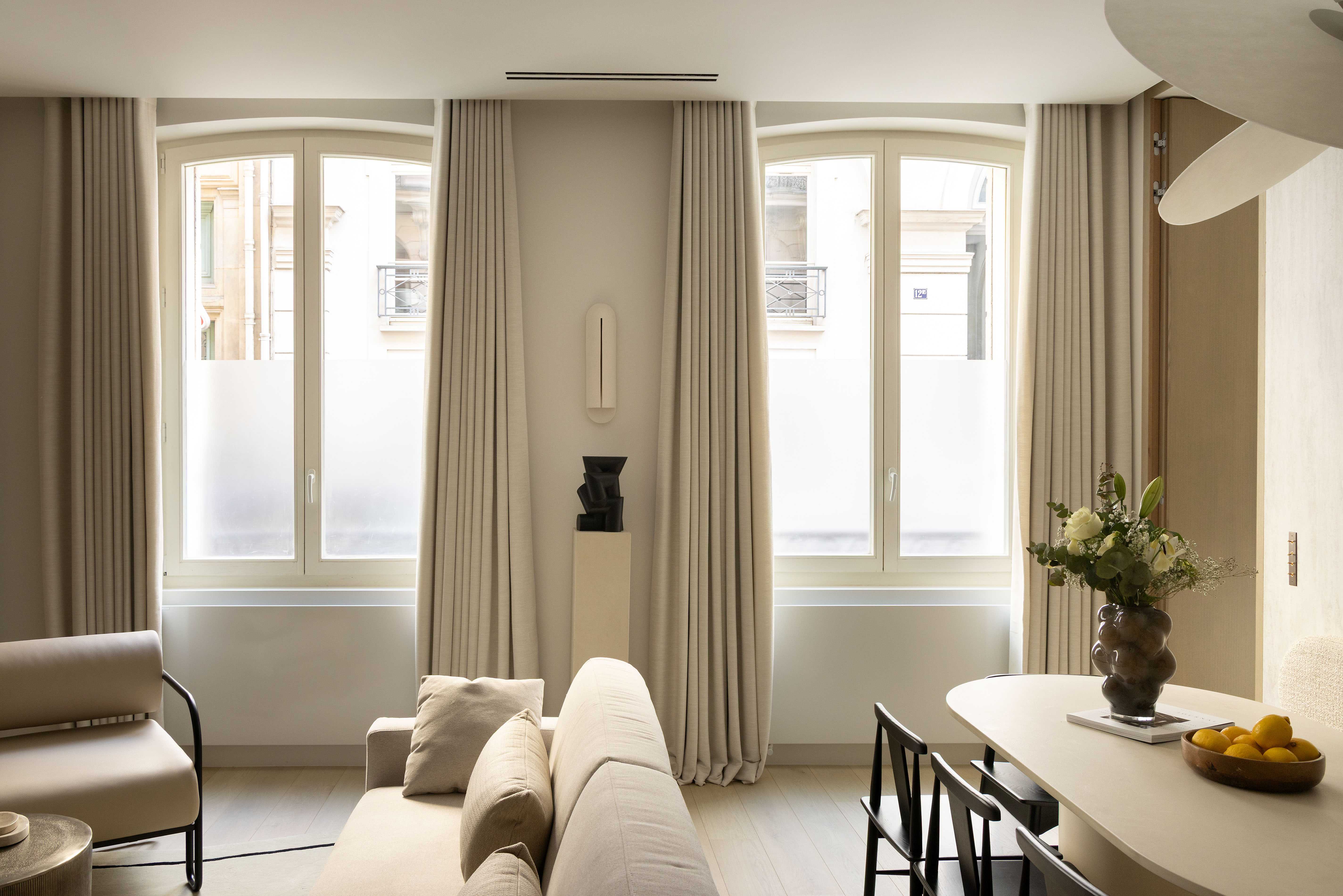 Salon lumineux avec canapé beige, grandes fenêtres cintrées, rideaux et table à manger