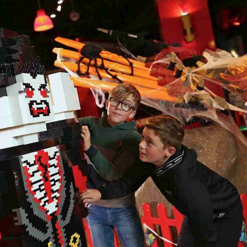 İki erkek çocuk, büyük bir LEGO vampir figürüyle etkileşimde bulunuyor. Arka plan, Cadılar Bayramı temalı bir düzenlemede örümcekler ve örümcek ağları ile dekore edilmiştir.