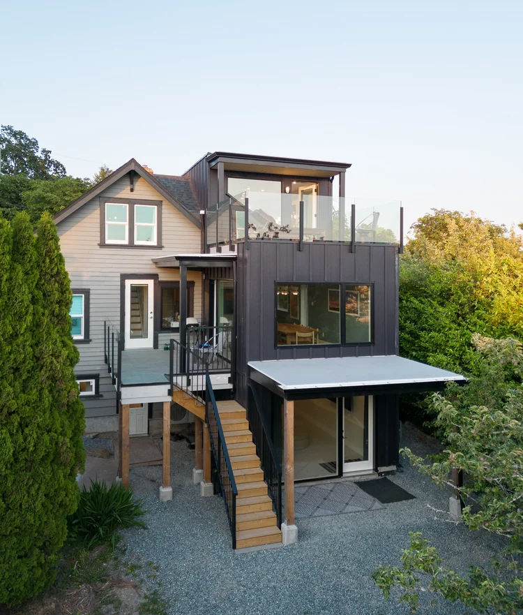 Modern two-story home with large glass windows, warmly lit interiors, and a balcony. Surrounded by trees and a manicured lawn at dusk. Cozy ambiance.