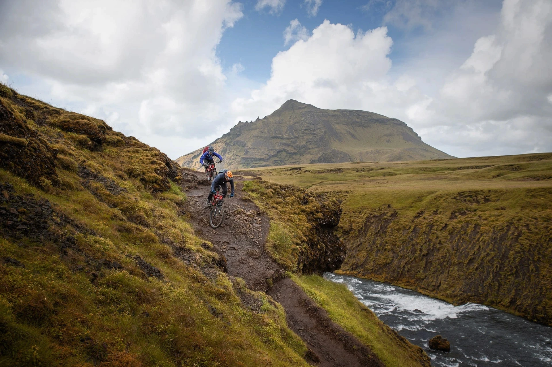 Two mountain bikers riding a narrow trail beside a river gorge.
