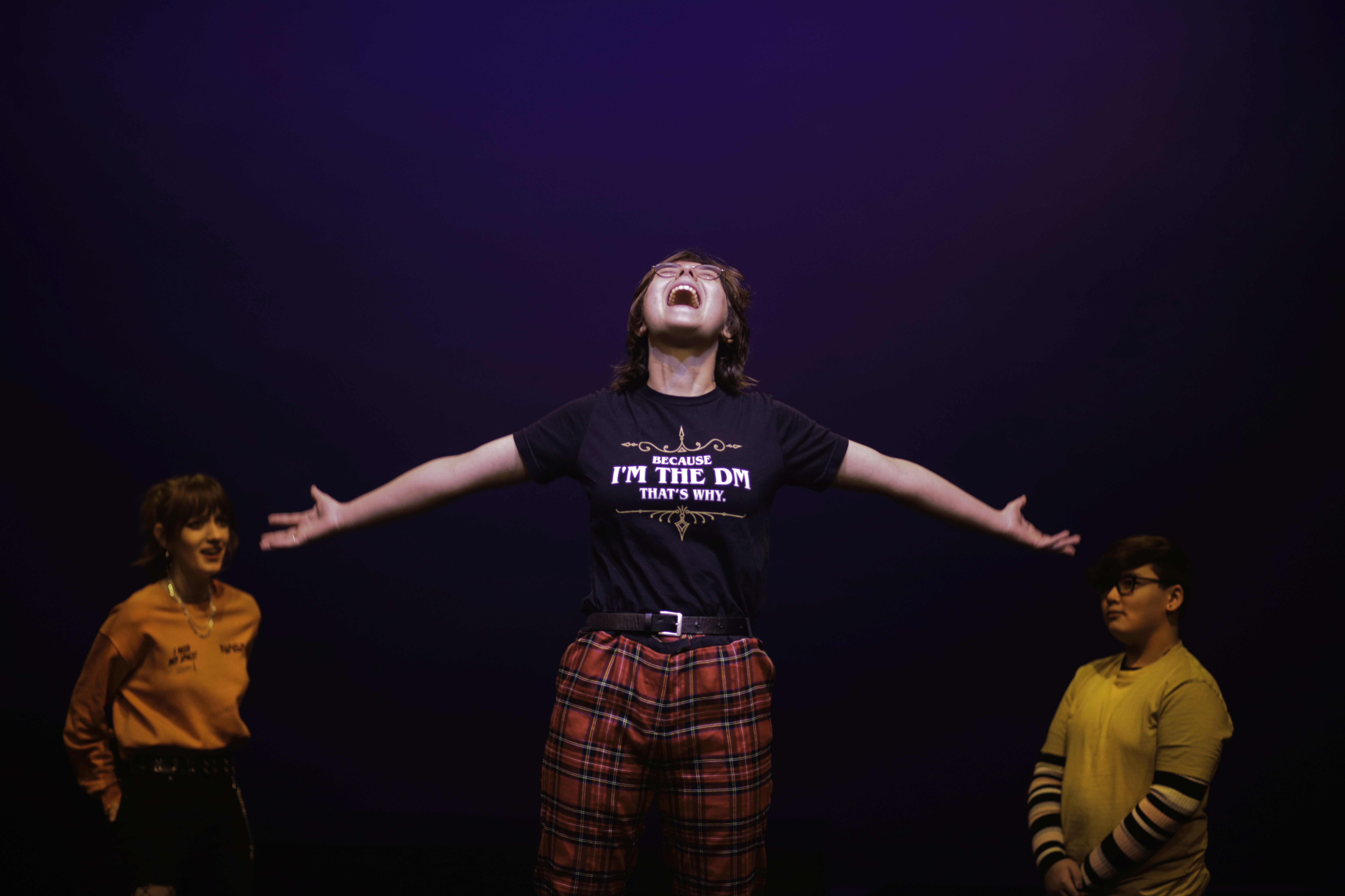 A teenager wearing red plaid pants and a black T-shirt that reads “Because I’m the DM, that’s why.” stands center stage with arms stretched wide, shouting passionately. Two other teens stand on either side, watching under purple stage lighting.