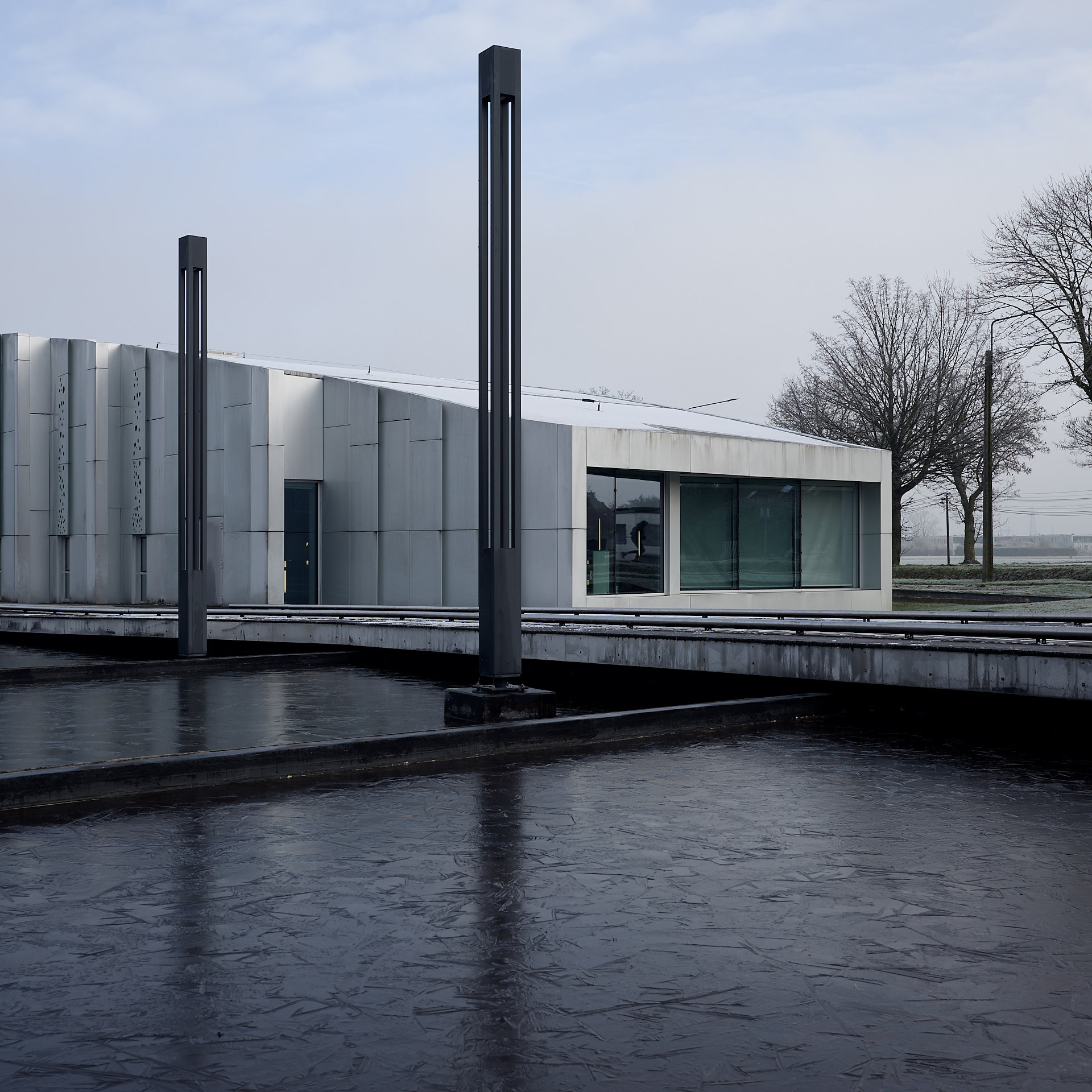 Photographie d'architecture extérieure du Centre Culturel de Lesquin : façade moderne blanche et rythmée se reflétant dans un bassin d'eau gelé sombre avec des luminaires verticaux graphiques.