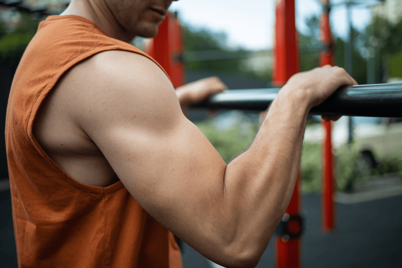 Close-up of a man’s arm and shoulder muscles as he performs a pull-up on outdoor bars.