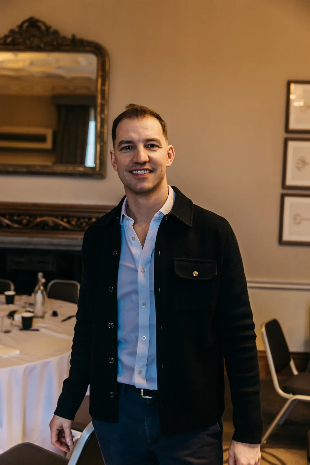 A person smiling and holding a drink in front of tables.