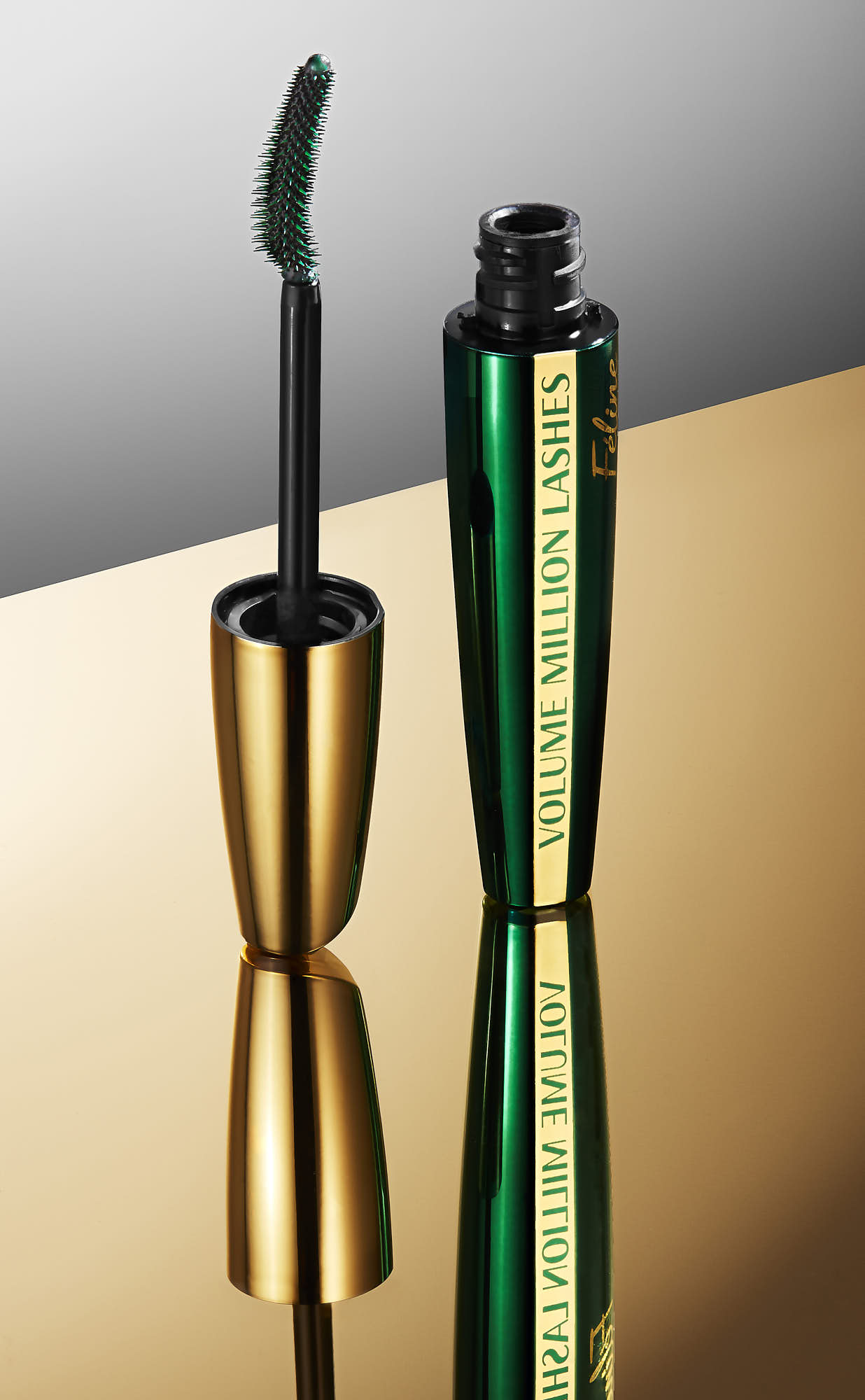 Two beauty tools are displayed: a metallic eyelash curler and a green mascara tube, reflecting on a shiny surface.