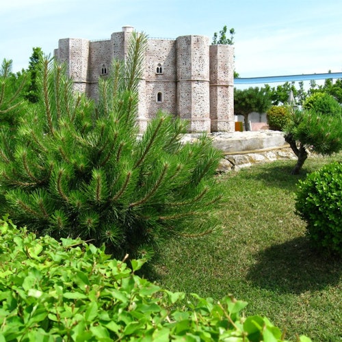 A miniature castle surrounded by lush greenery, bushes, and small pine trees under a clear blue sky.