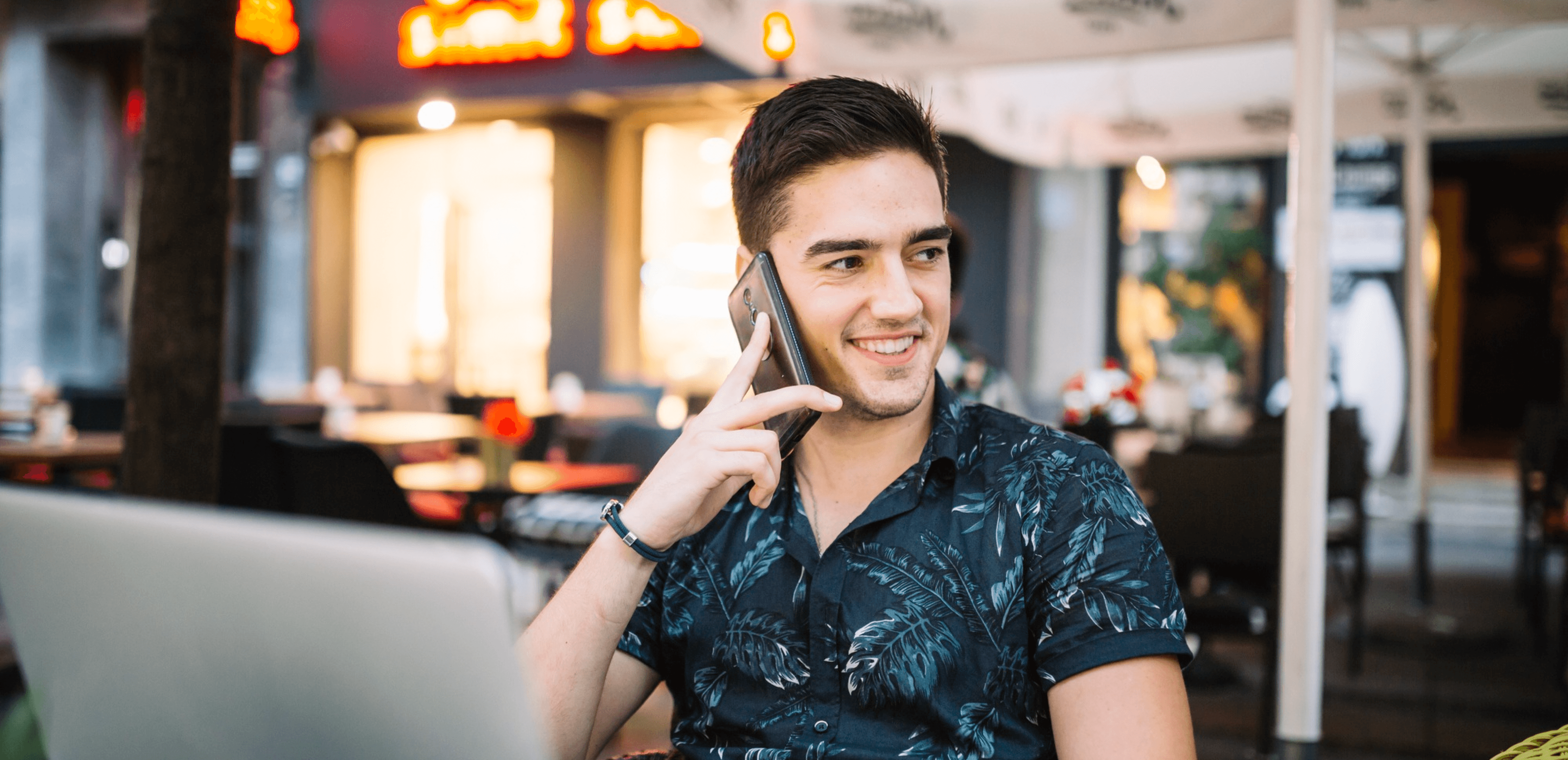 A young man sits at an outdoor cafe table, smiling while holding a phone to his ear, with an open laptop in front of him and warm ambient lighting from nearby buildings enhancing the scene.