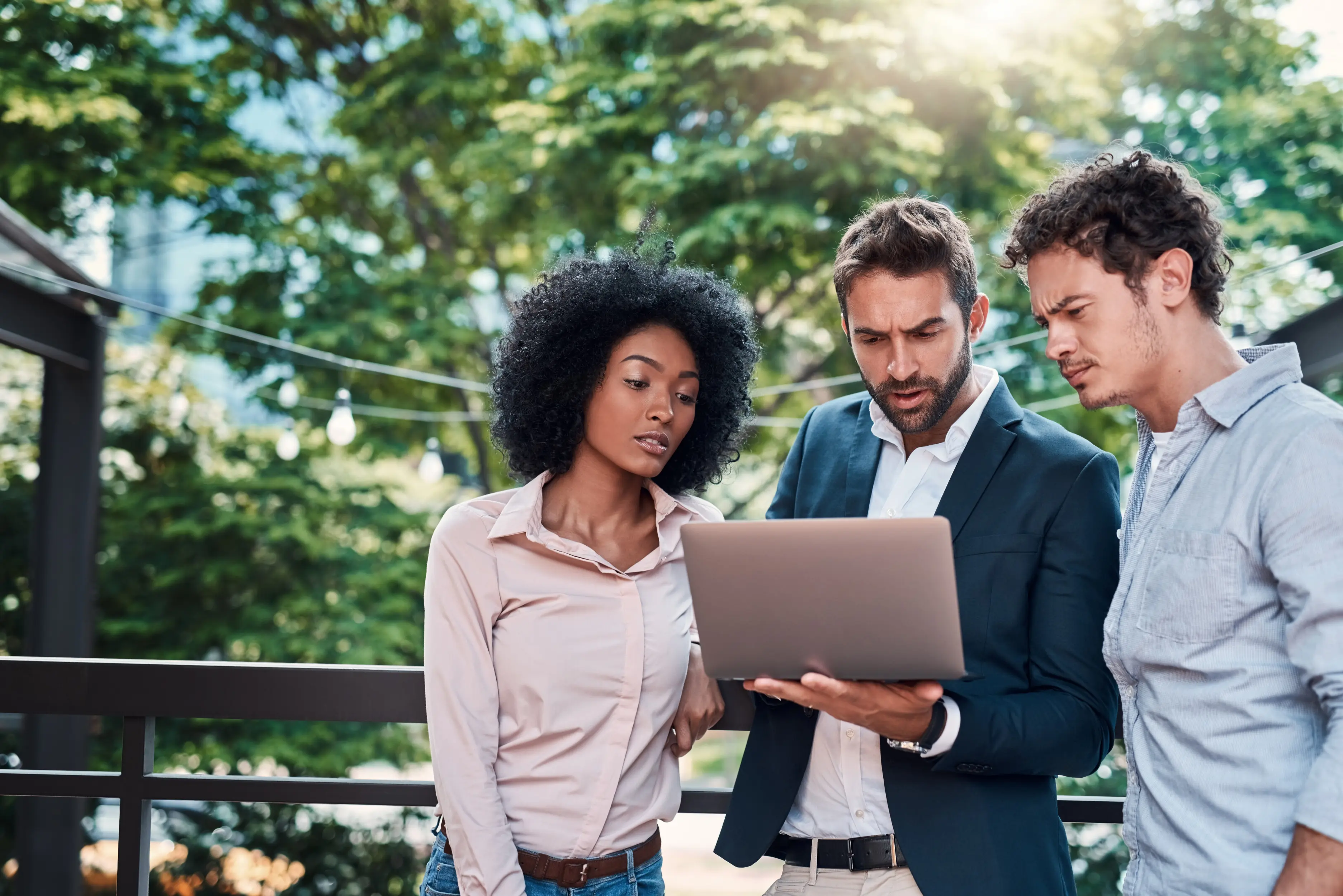 A woman on the left wearing a pink shirt, a man in the middle wearing a suit holding a laptop and a man on the right wearing a blue shirt. They are all looking ath the open laptop screen