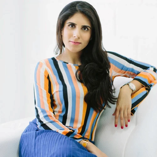A woman with long hair poses confidently, wearing a colorful striped top and sitting against a white background.