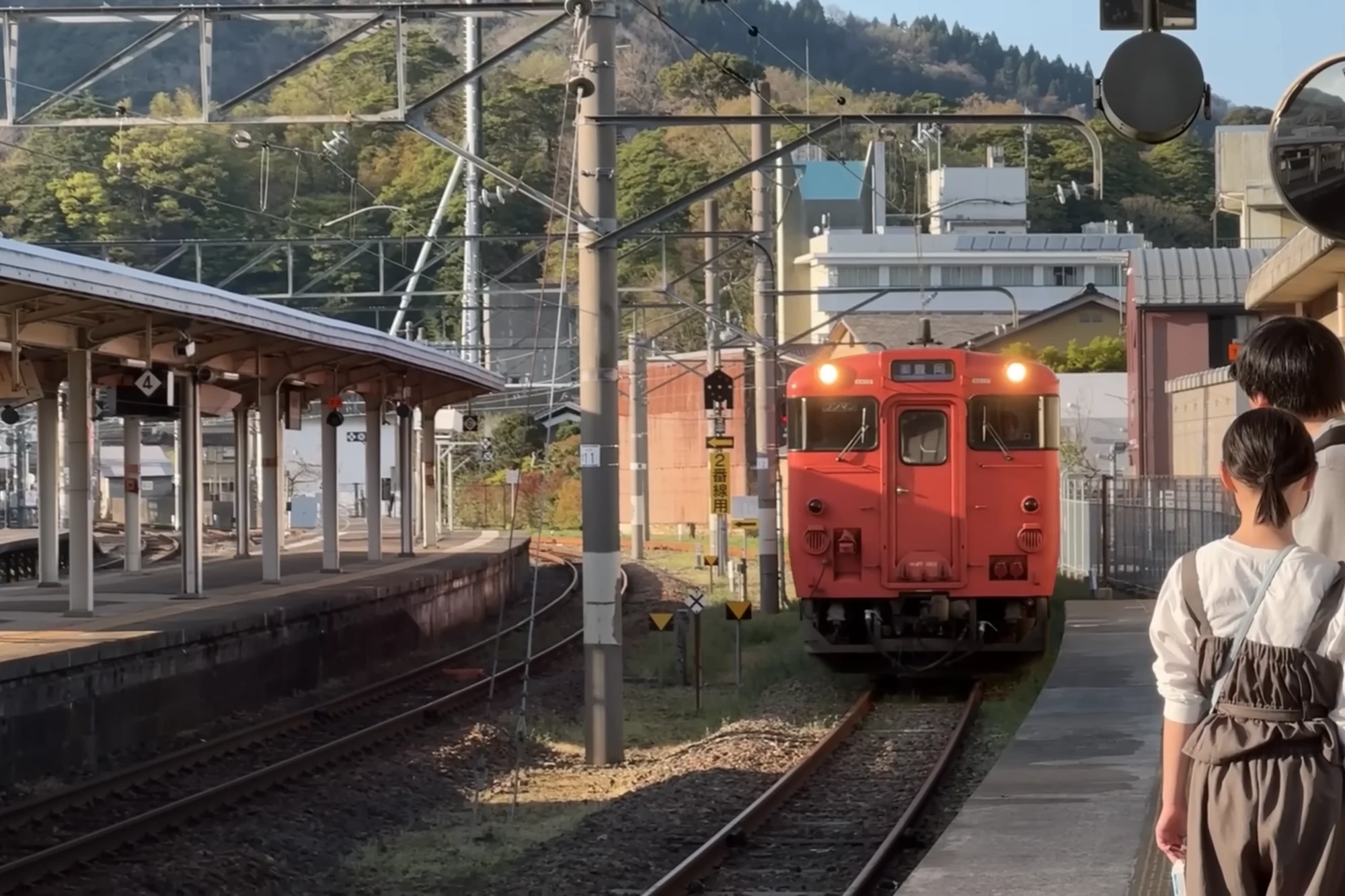 A red two-car railcar drawing into a rural platform with two people waiting on the edge.