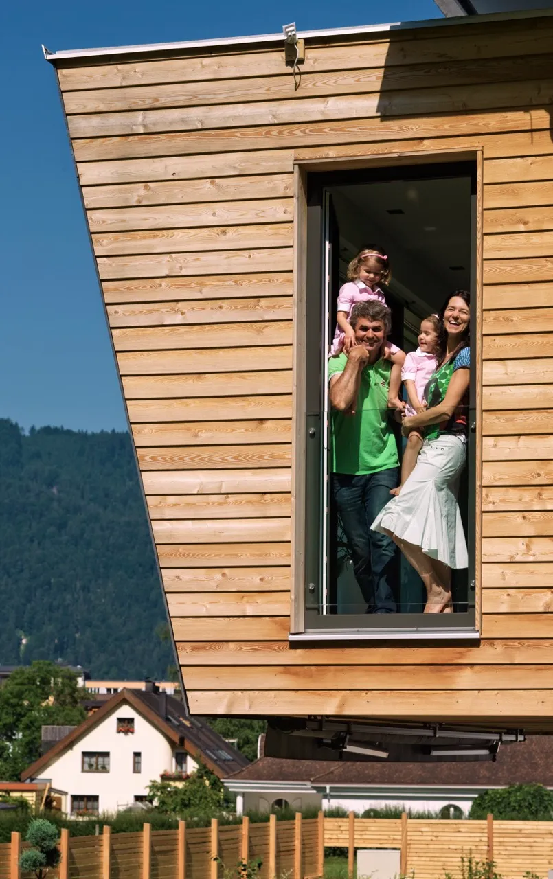 Familie mit zwei Kindern steht lächelnd am Fenster eines Holzhauses, eine Tochter auf den Schultern des Vaters.