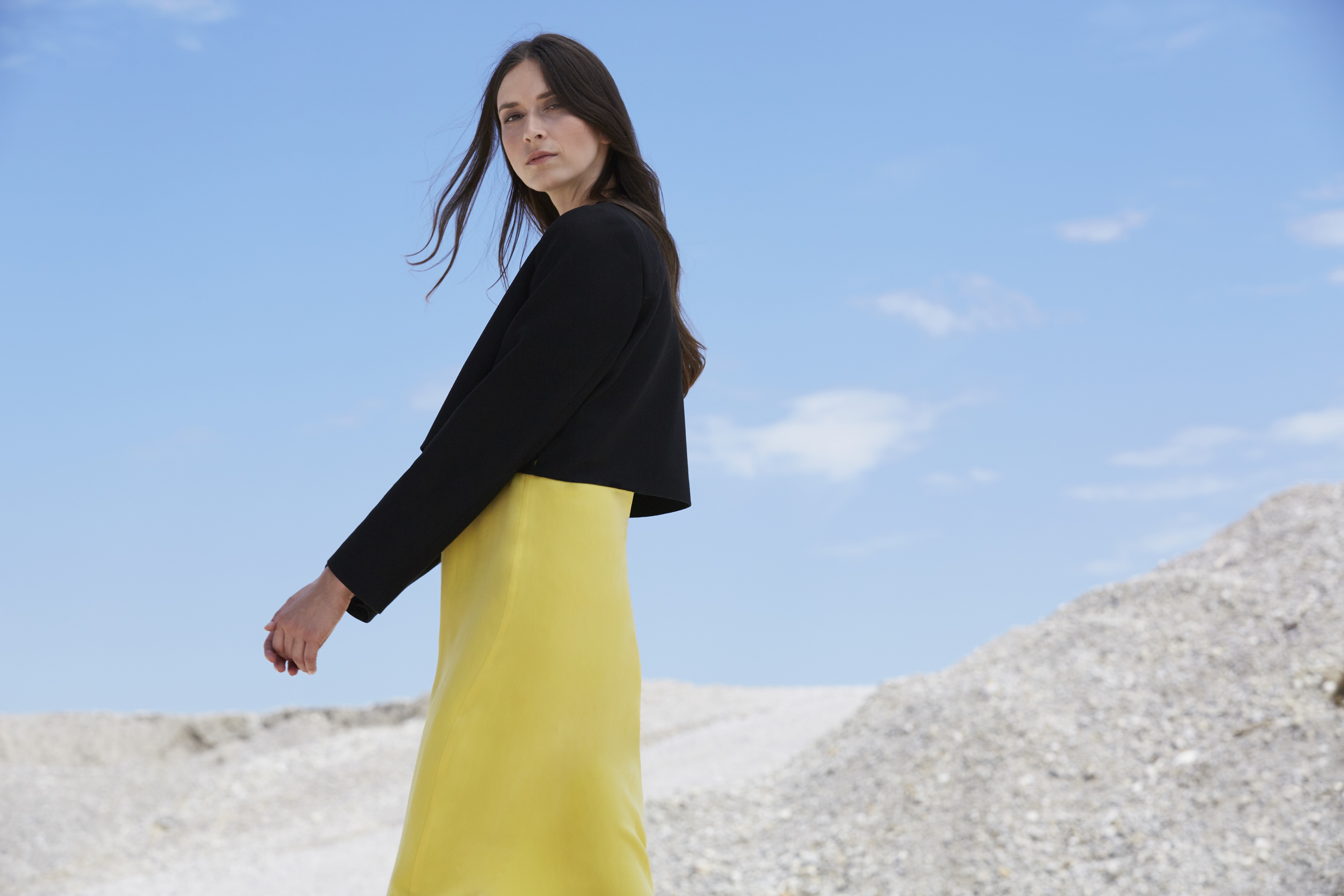 A woman in a yellow dress and black jacket stands against a backdrop of white rocky hills under a clear blue sky.