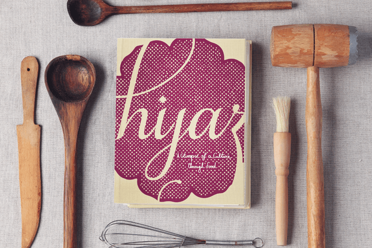 A cookbook titled “Hijab: A Clump of a Culture Through Food” is centered on a light gray surface. Surrounding the book are various wooden kitchen utensils: a wooden spoon, a wooden knife, a wooden spatula, a wooden mallet, a wooden brush, and a metal whisk.