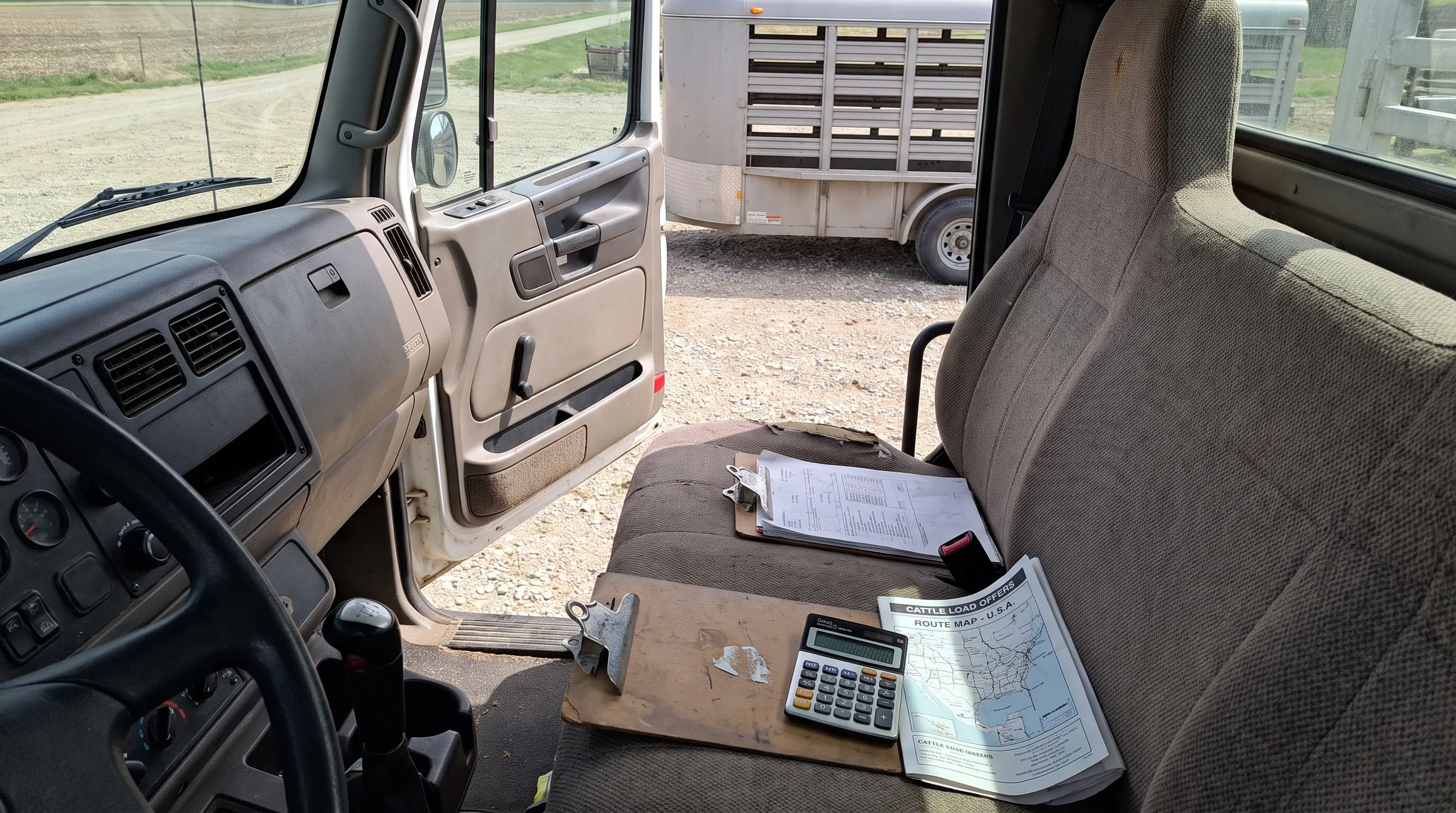 A clipboard, calculator and route map sit on a truck’s passenger seat as a cattle trailer is visible through the open cab door, ready for comparing load offers.