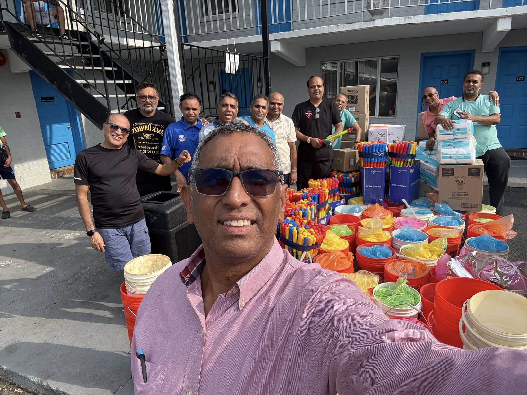 Two smiling people outdoors, one in a pink jacket holding a white phone, the other in blue with a buzz cut holding a blue phone, under a clear sky.