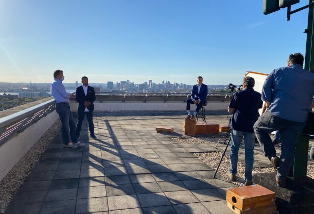 Three men in business attire film an interview on a rooftop with a city skyline in the background, while two crew members operate cameras and lighting equipment.