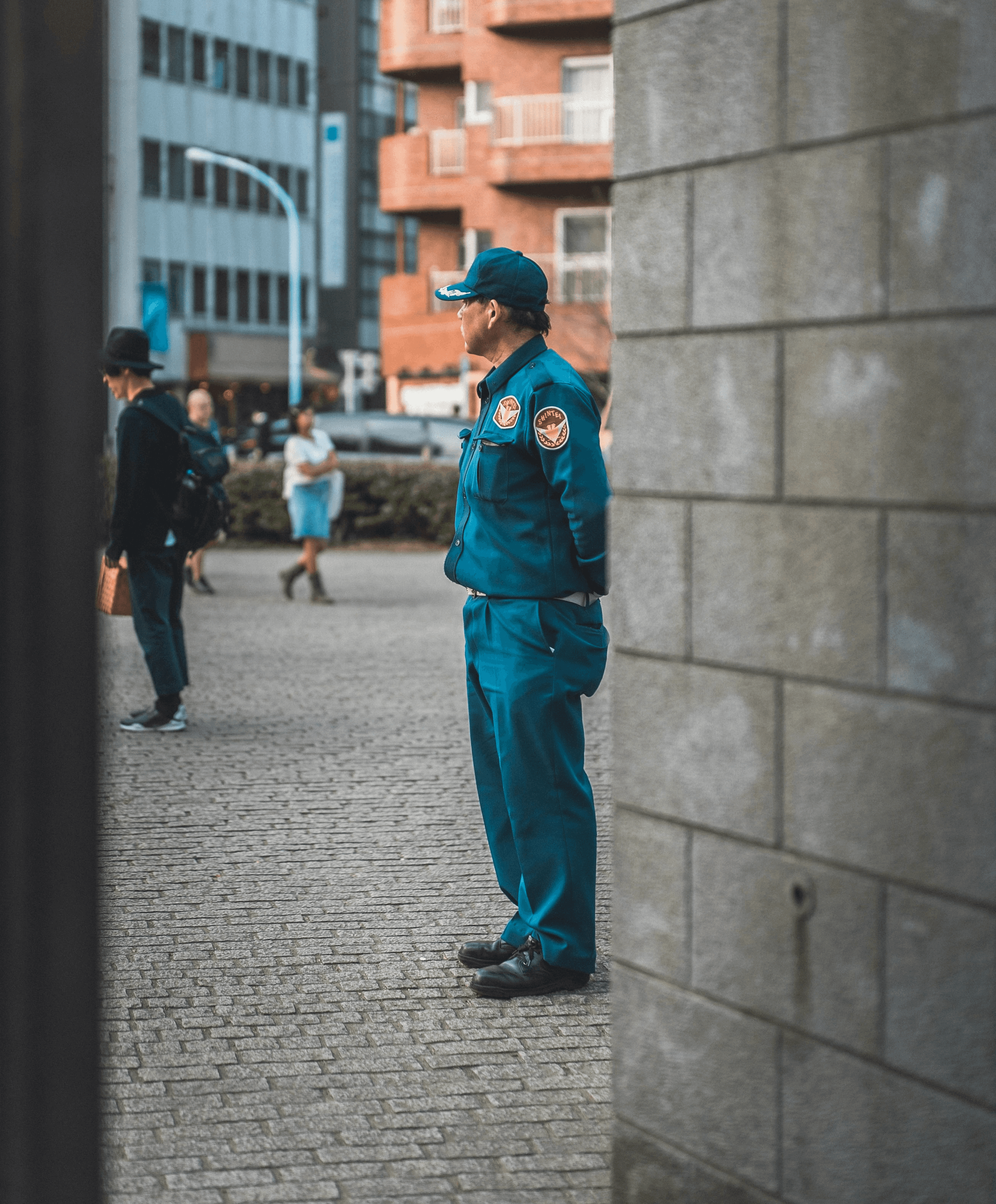A uniformed officer stands guard next to a stone wall, with people in the background blurred out.