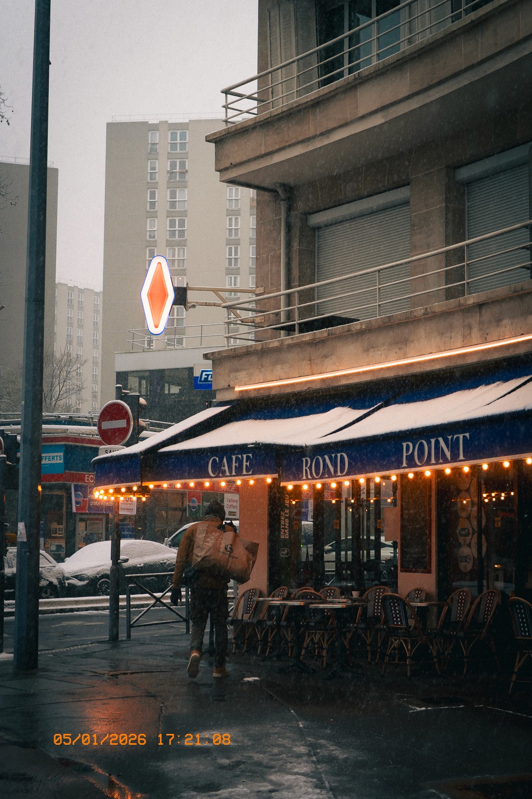 Rue enneigée à Issy avec le café 'Café Rond Point', une personne marchant avec un sac, des immeubles résidentiels en arrière-plan, un panneau lumineux en forme de losange et des voitures recouvertes de neige sous un ciel gris.