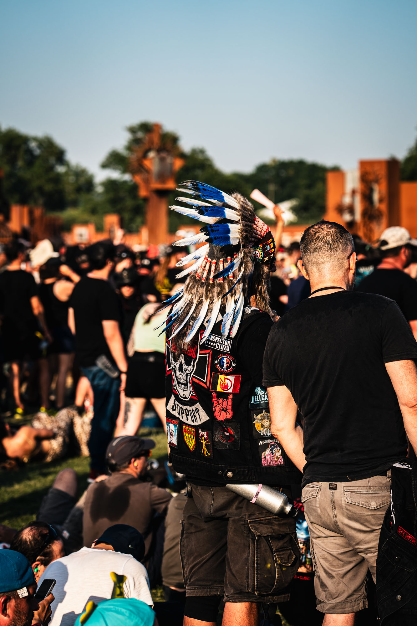 Festivalier de dos coiffé d'une parure de plumes bleues et blanches, foule assise au soleil