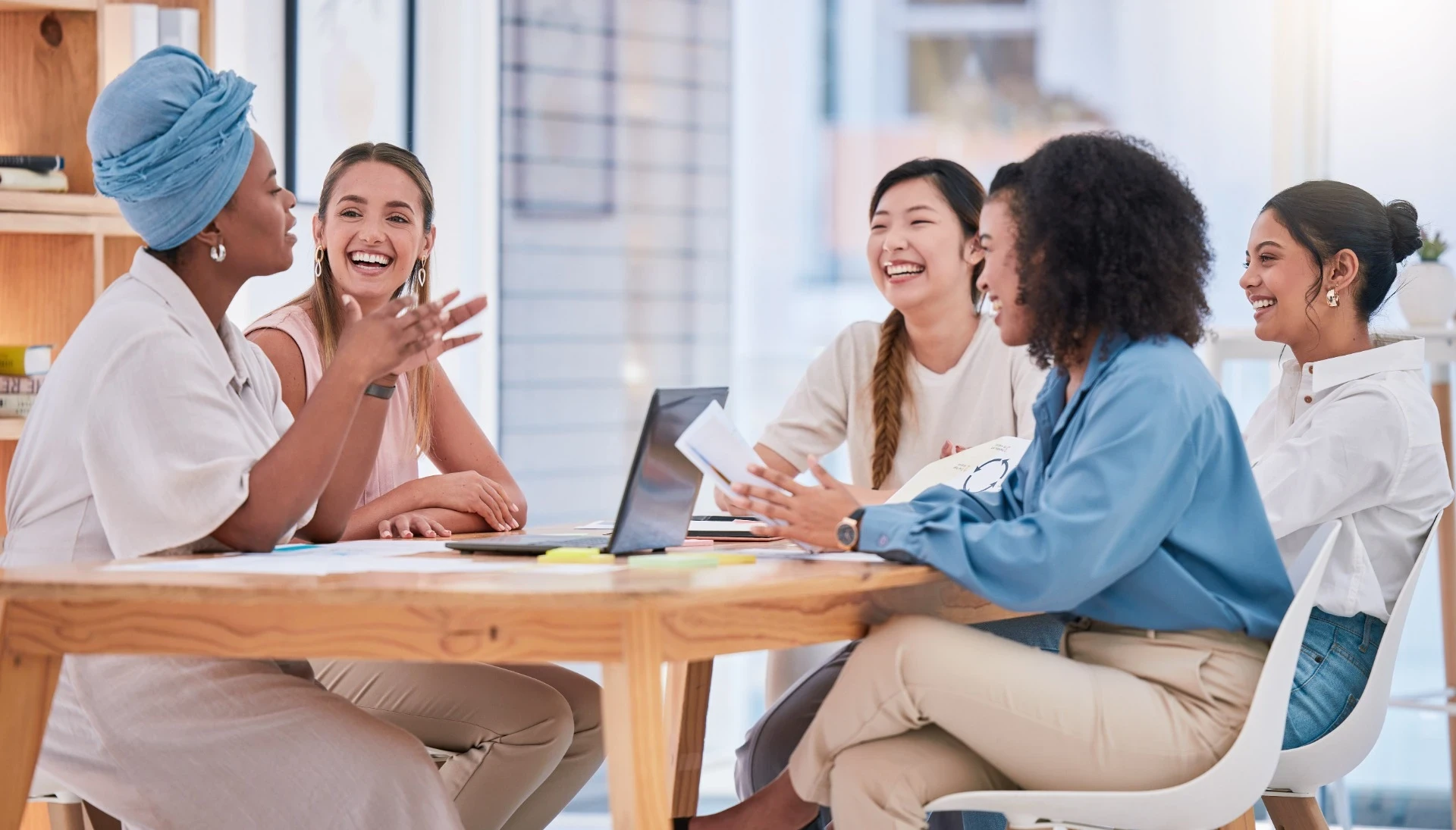 A imagem mostra um grupo de cinco mulheres reunidas ao redor de uma mesa, envolvidas em uma conversa animada e alegre. Todas estão sorrindo, demonstrando um ambiente descontraído e colaborativo. Elas estão em um espaço moderno e iluminado, provavelmente um ambiente de trabalho ou uma reunião informal. Sobre a mesa há um laptop aberto e documentos, sugerindo que estão discutindo ideias ou projetos. As mulheres estão vestidas de forma casual, e uma delas usa um turbante azul claro, acrescentando um toque de estilo pessoal ao conjunto. A interação entre elas transmite um espírito de colaboração, diversidade e energia positiva.