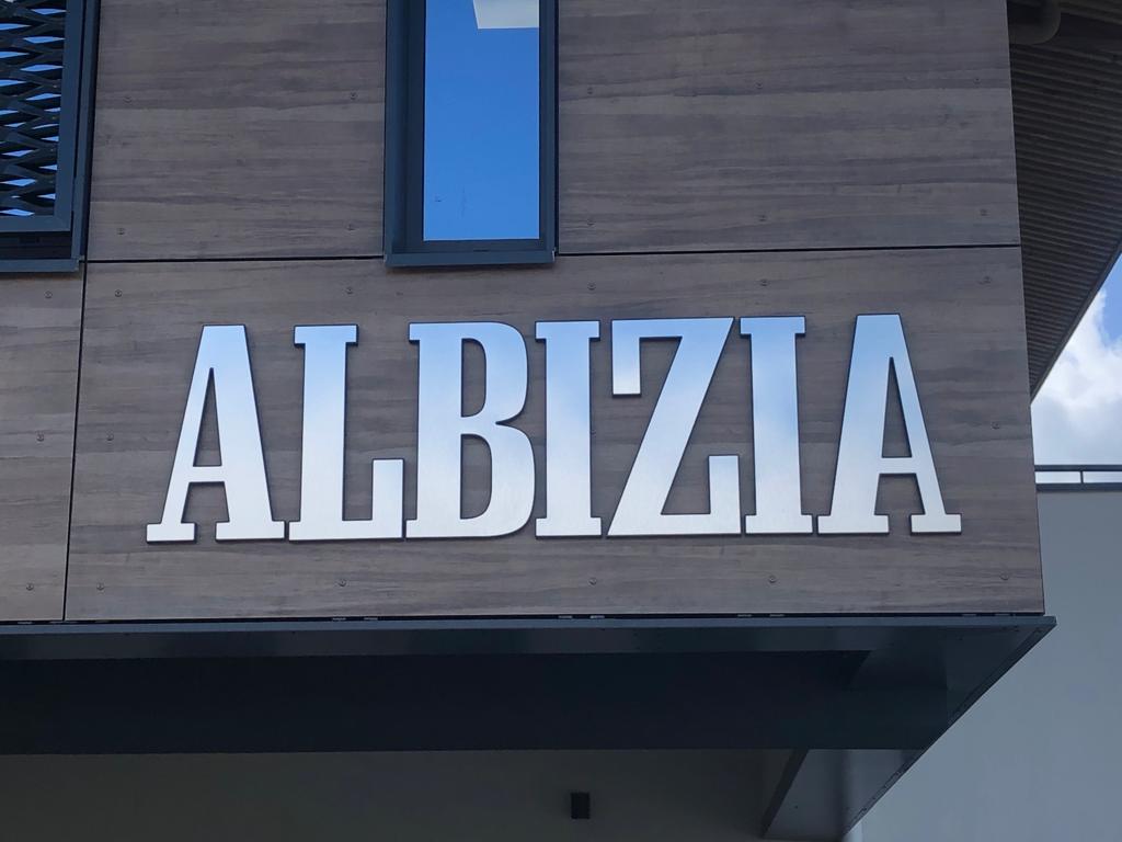 Une enseigne en lettres argentées formant le mot « ALBIZIA » sur une façade en bois. Une fenêtre au-dessus reflète un ciel bleu. Ambiance moderne et sophistiquée.