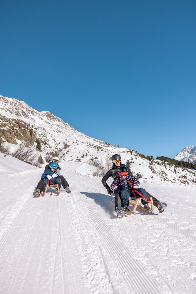Zwei Personen mit Helmen und Winterkleidung rodeln unter einem klaren blauen Himmel einen schneebedeckten Berghang hinunter, umgeben von schneebedeckten Gipfeln im Ötztal.