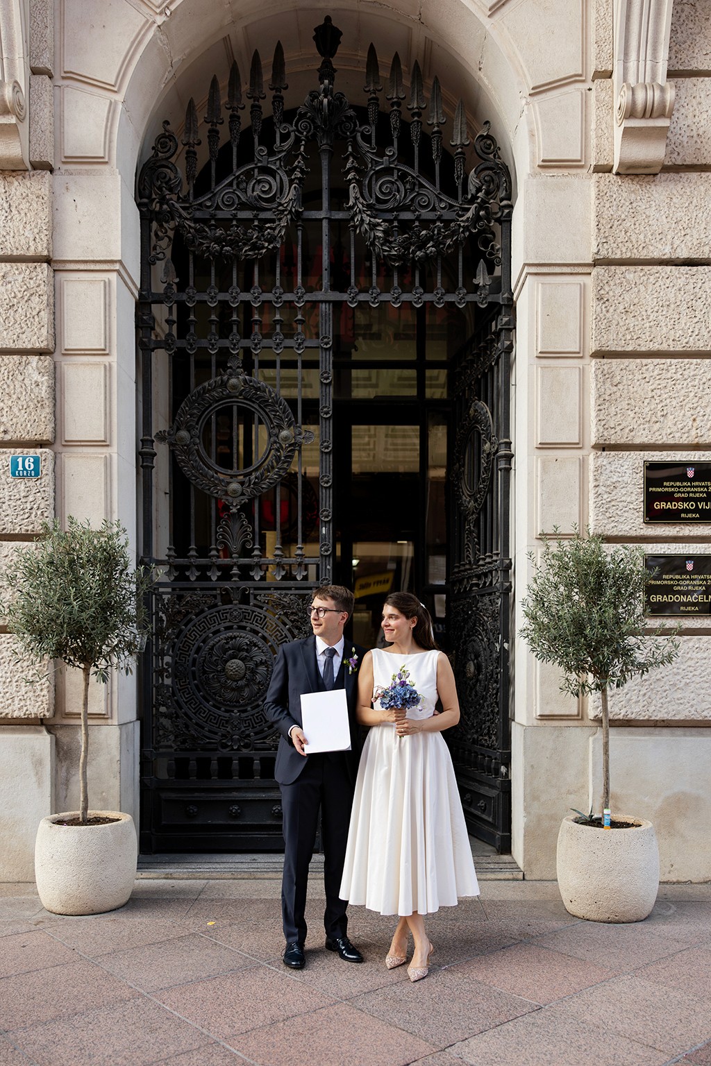 Just married couple posing for a photo in front of two metal gates, doors to the city hall, holding a marriage certificate and a bouquet of flowers